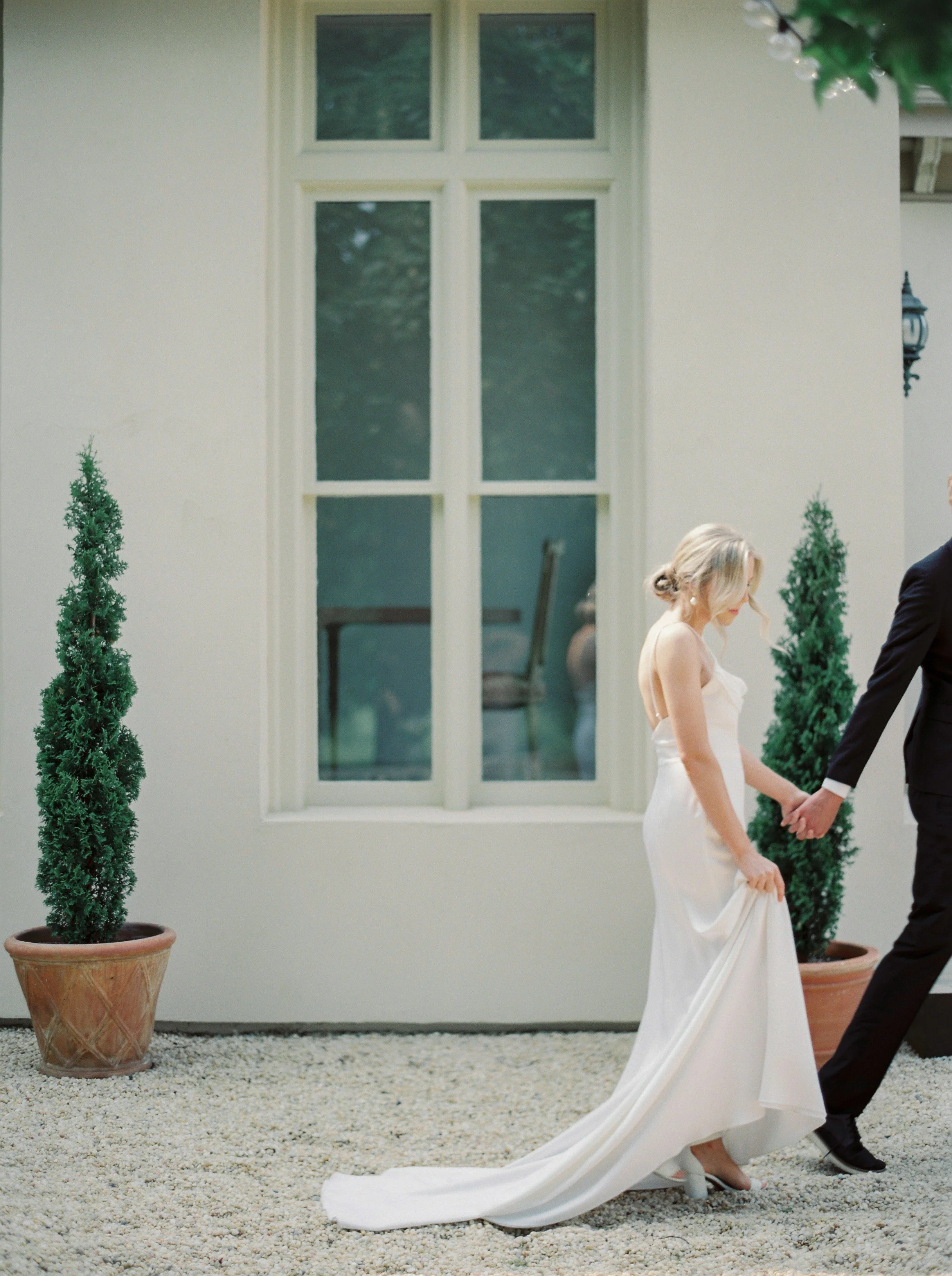 Bride in a white wedding gown holding hands with groom in a black suit outside a building, with potted trees and a window in the background.