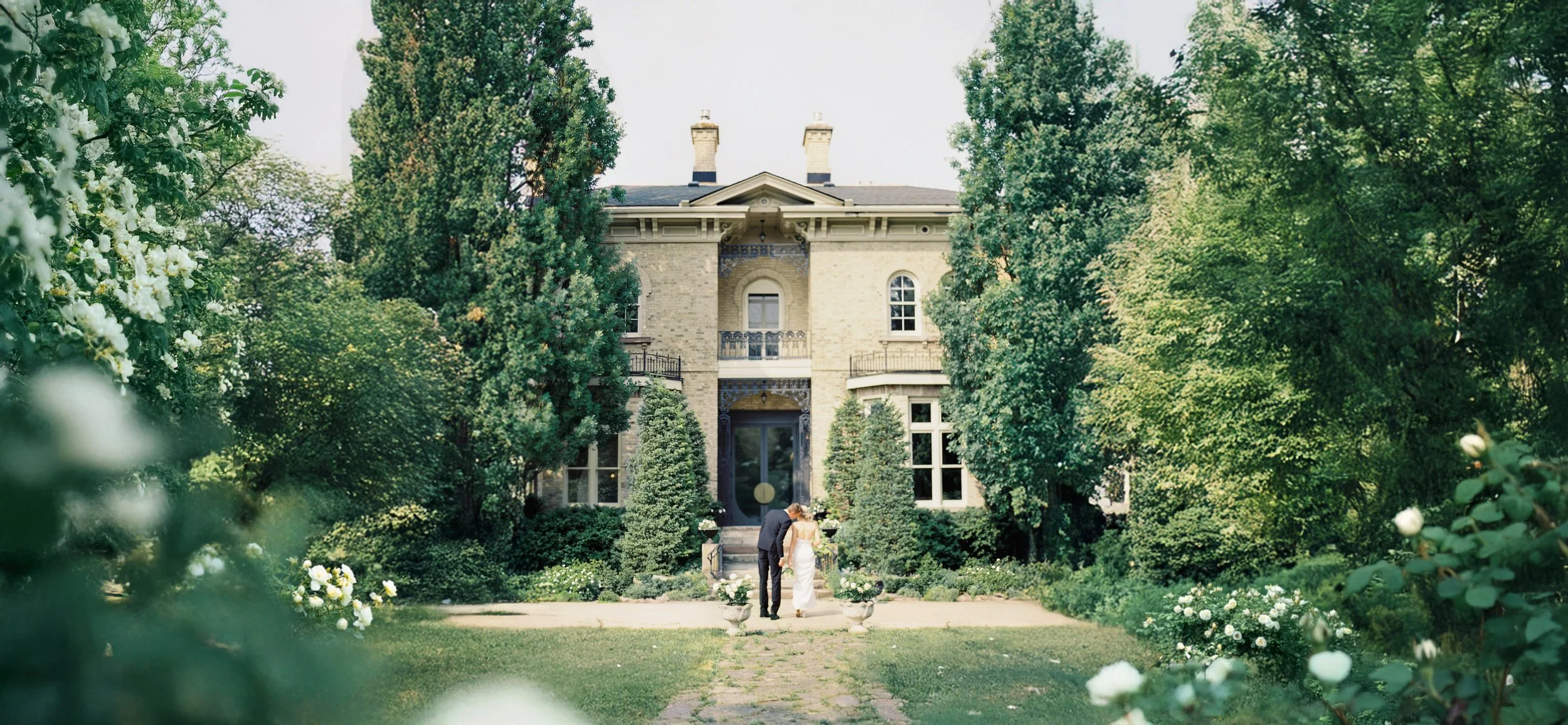 A couple in wedding attire walking hand in hand on a garden path in front of a large, historic mansion surrounded by lush green trees and flowering bushes.