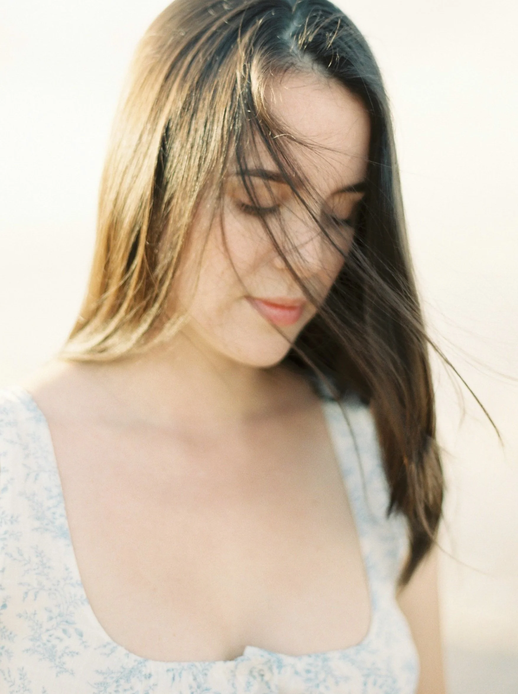 A young woman with long brown hair, wearing a light-colored top with a floral pattern, looking down with a gentle expression.