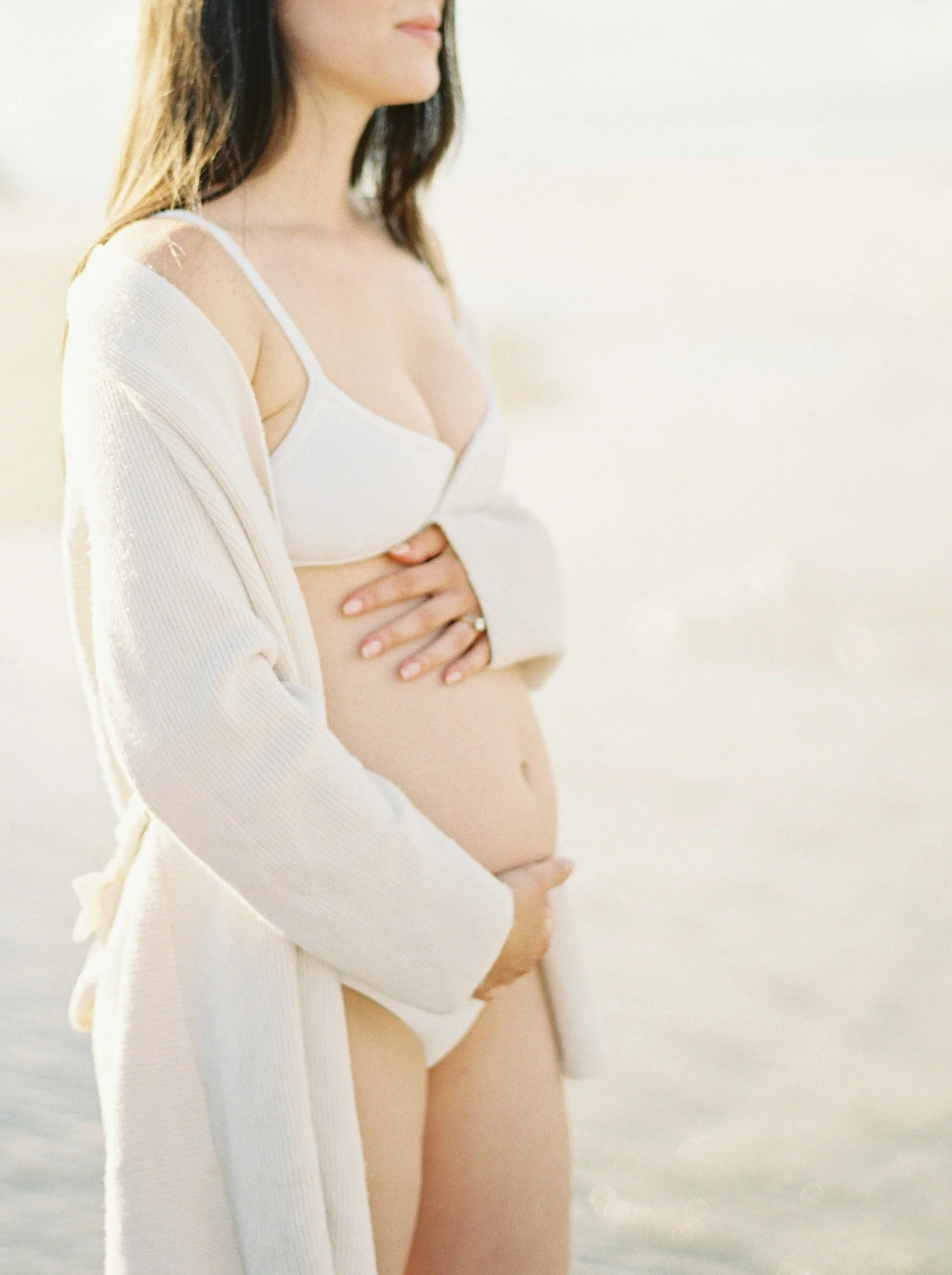 A woman at the beach wearing a white bra and a cream-colored cardigan, holding her pregnant belly with one hand and placing the other on her chest, with soft lighting and an out-of-focus ocean background.