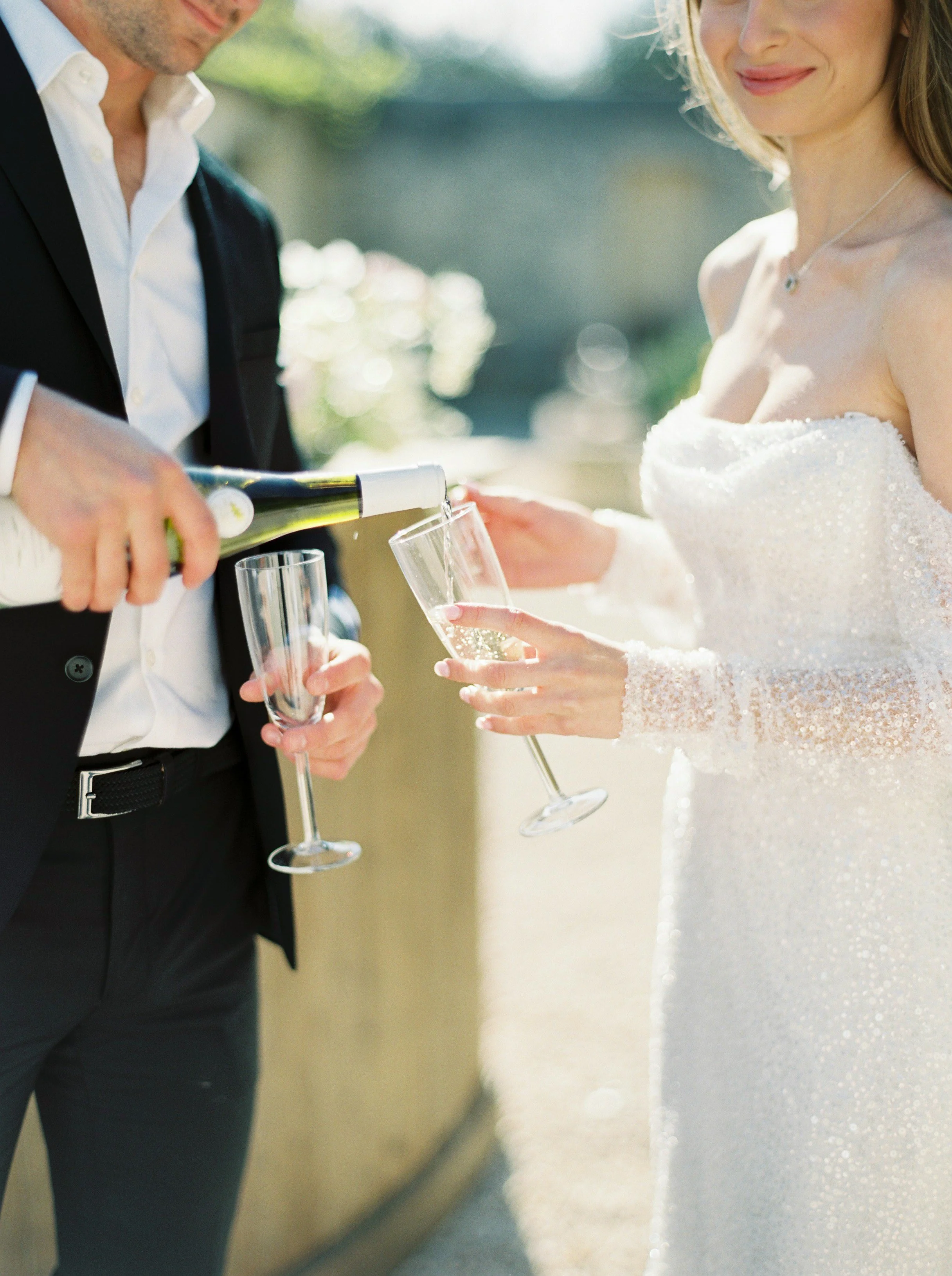 A bride and groom at their wedding reception, with the groom pouring champagne into the bride's glass outdoors.