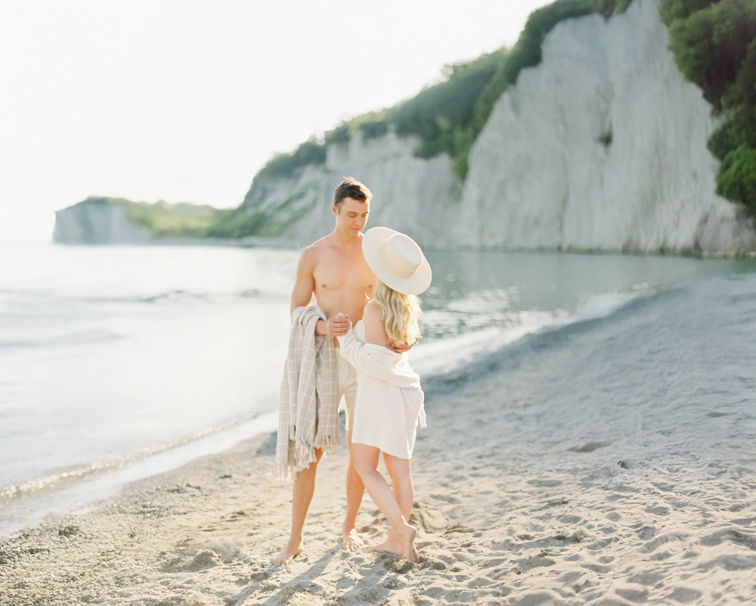 A young couple dressed in beachwear standing on a sandy beach by the water, with a large white cliff and greenery in the background, during daytime.