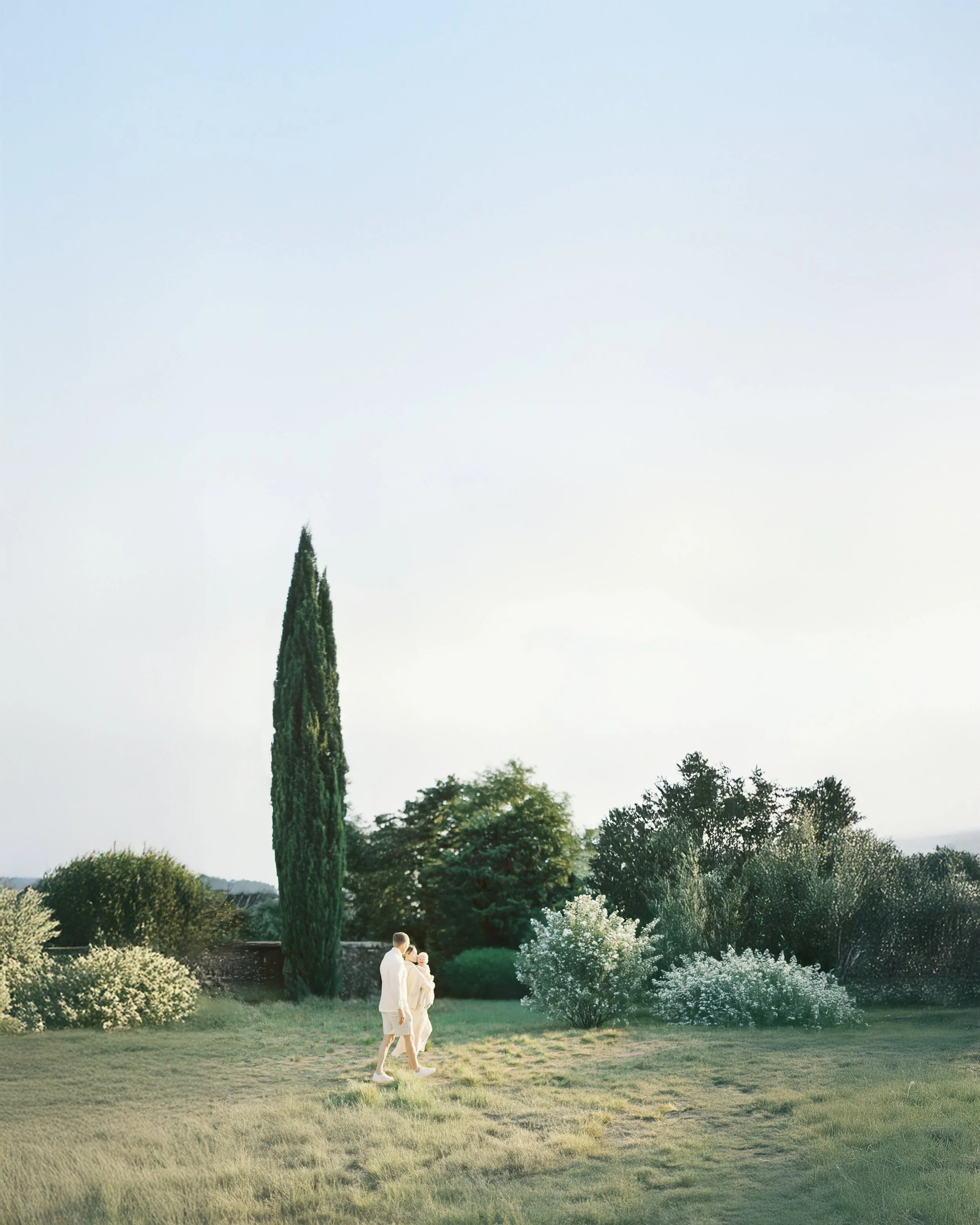 A couple walking in a lush green garden with trees and bushes, under a clear sky.