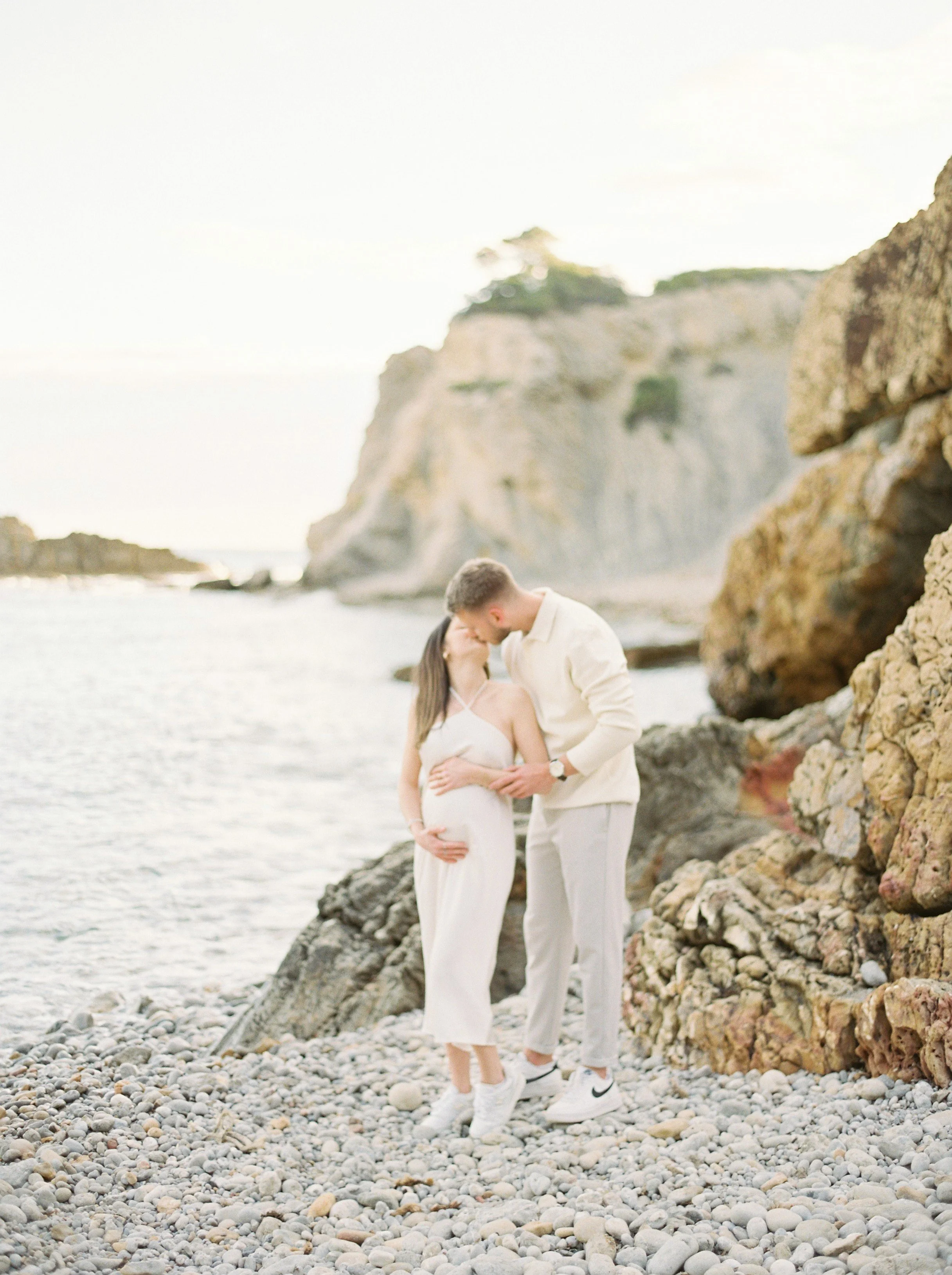 A couple standing on a rocky beach, with cliffs in the background, sharing a kiss.