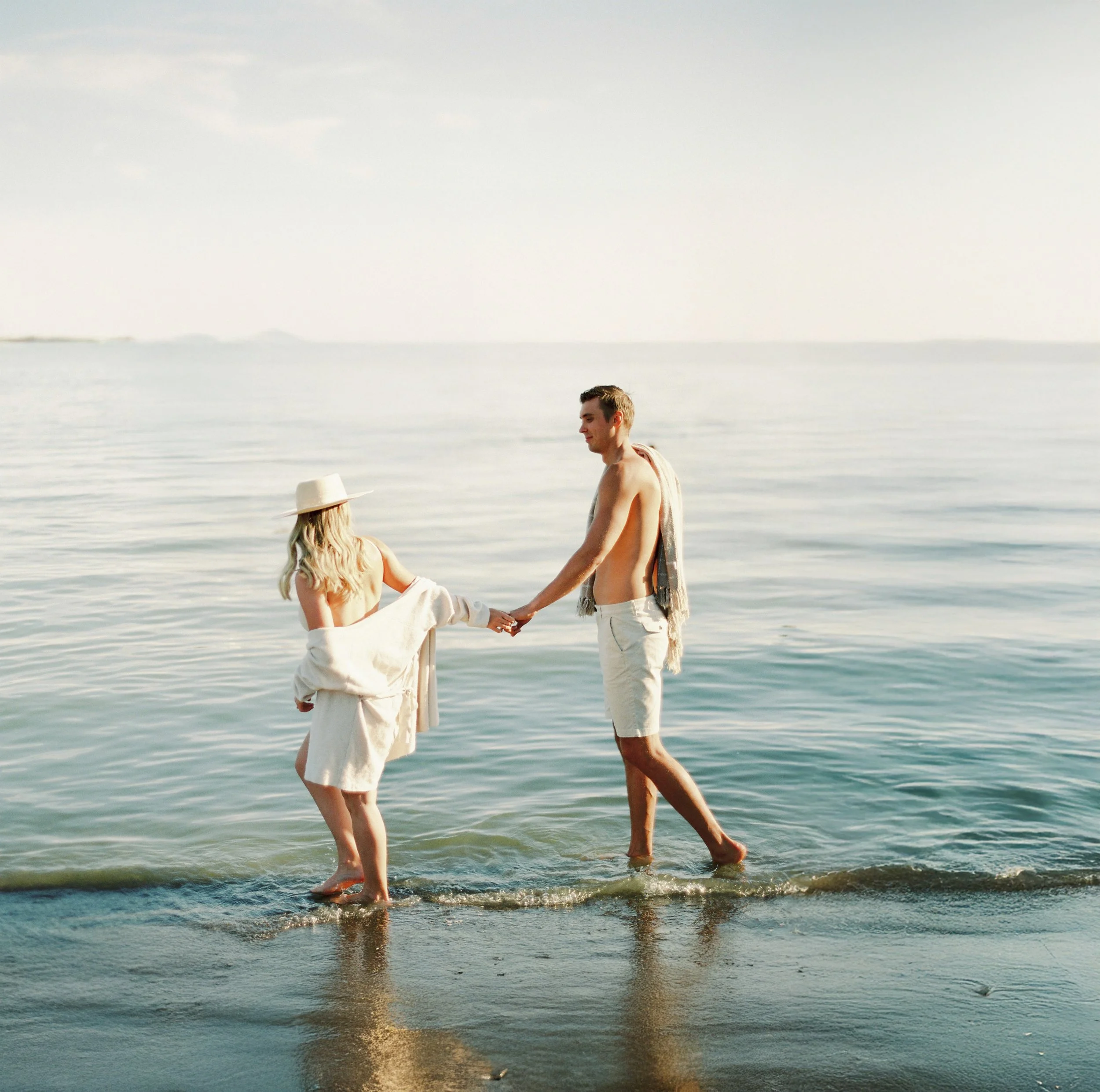 A man and woman holding hands and standing in shallow ocean water on a beach.