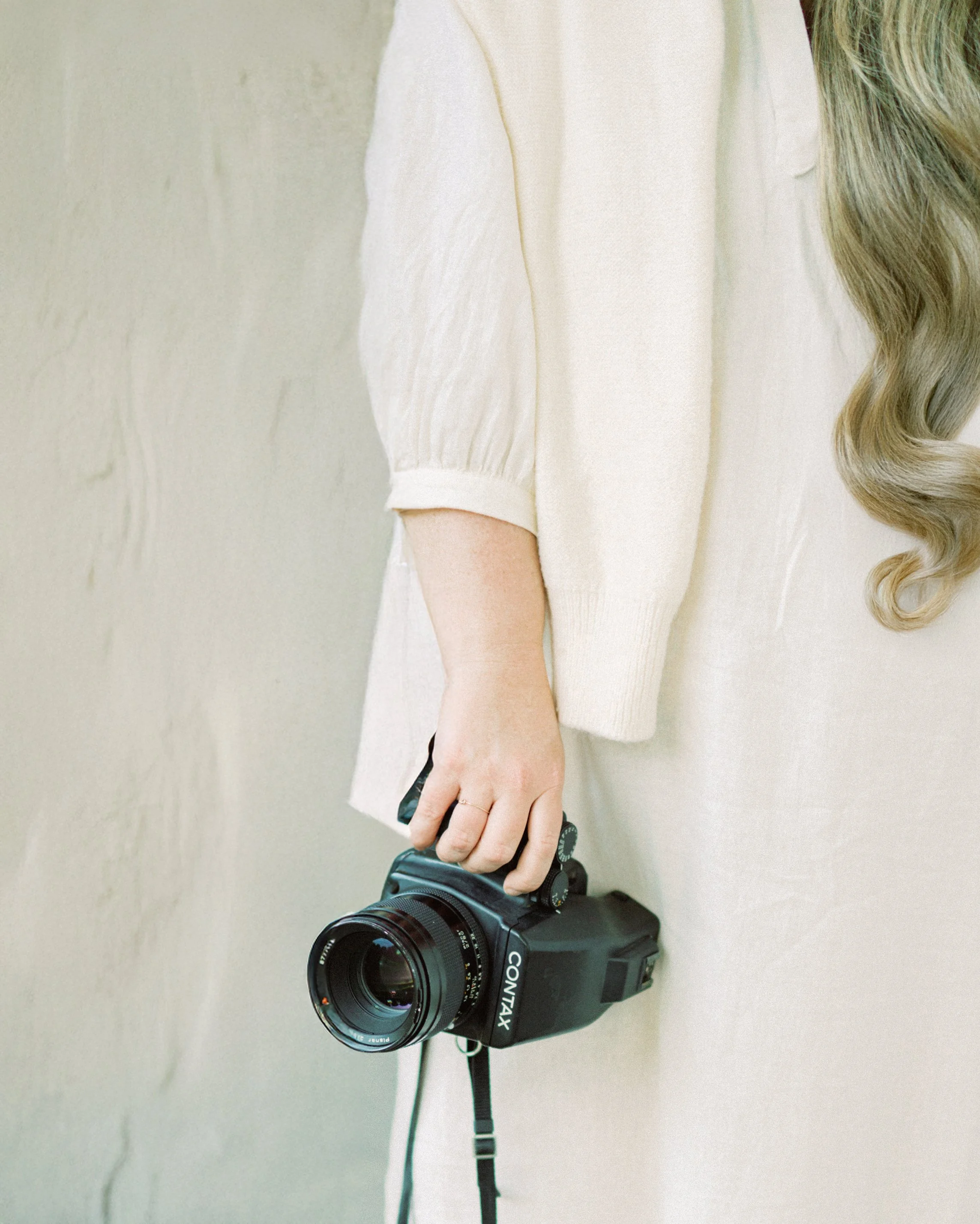 A person with long blonde hair wearing a light-colored shirt holding a black Canon camera by the side.