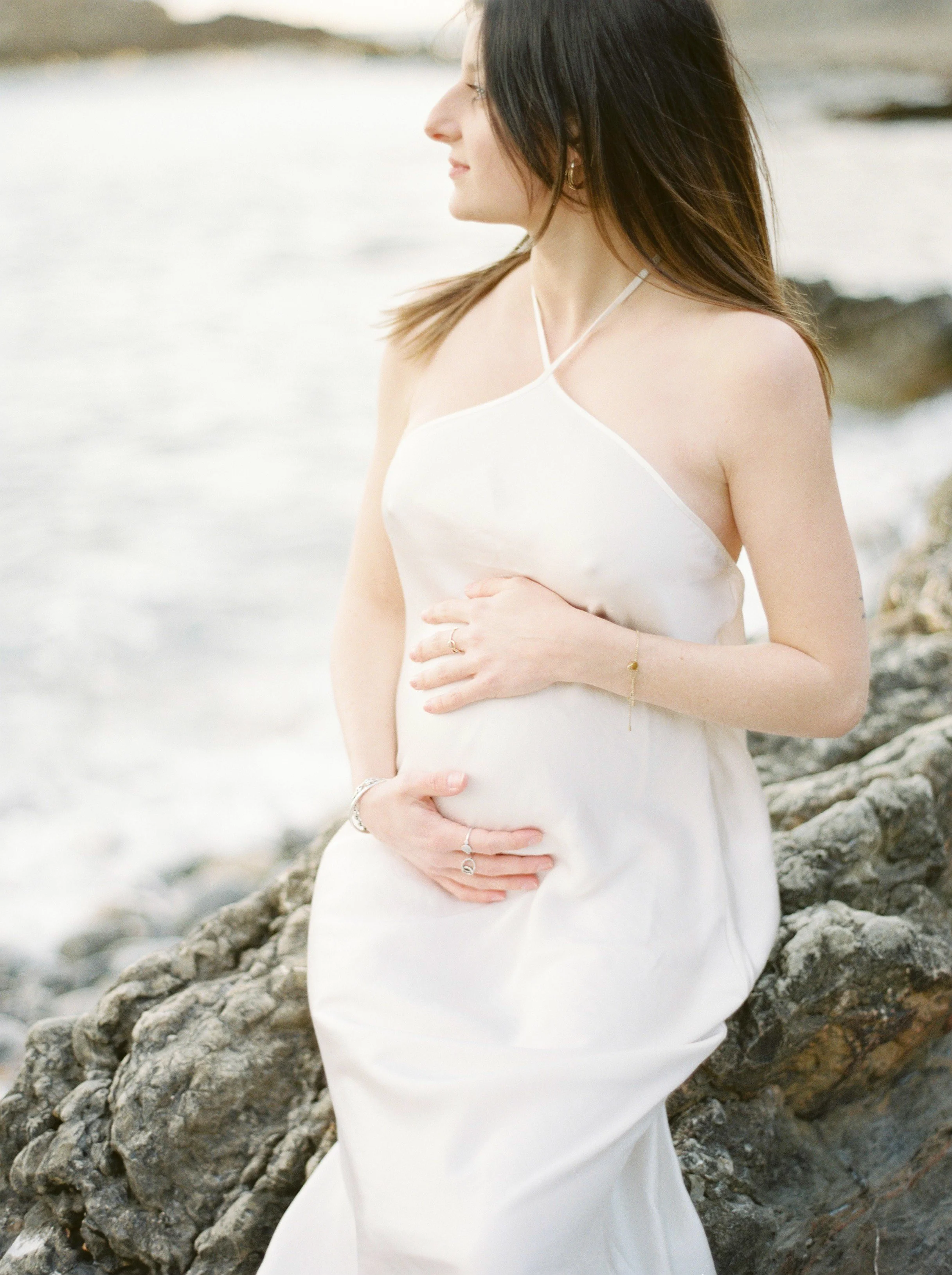 A pregnant woman wearing a white dress sits on rocks by the ocean during daytime, holding her belly with both hands and looking to the side.