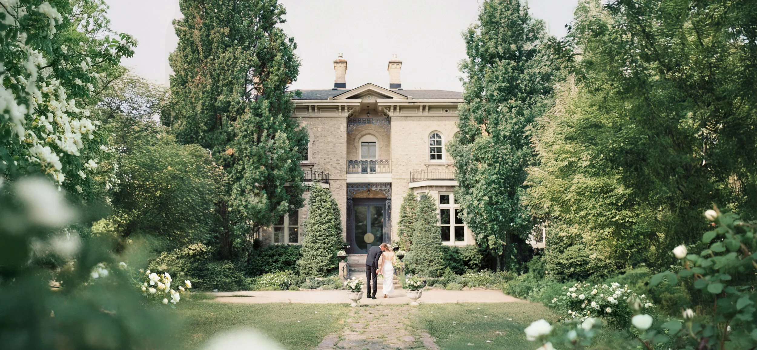 A bride and groom holding hands and leaning their foreheads together in front of a large, historic mansion surrounded by lush greenery and trees, with white flowers in the foreground.