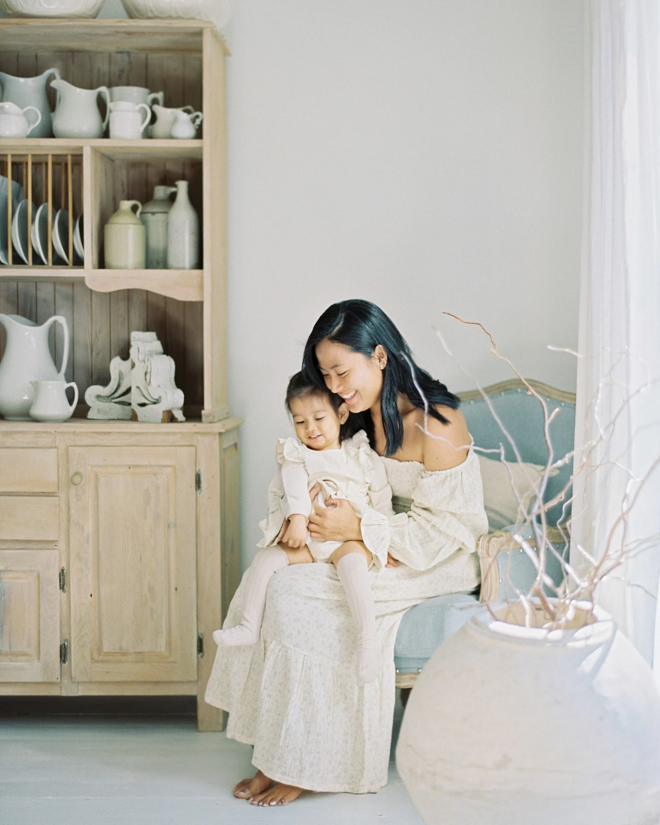 A woman sitting on a vintage armchair with a young girl on her lap, smiling and sharing a happy moment indoors near a blue and white vase with decorative branches, with a wooden cabinet filled with white and pastel ceramics in the background.