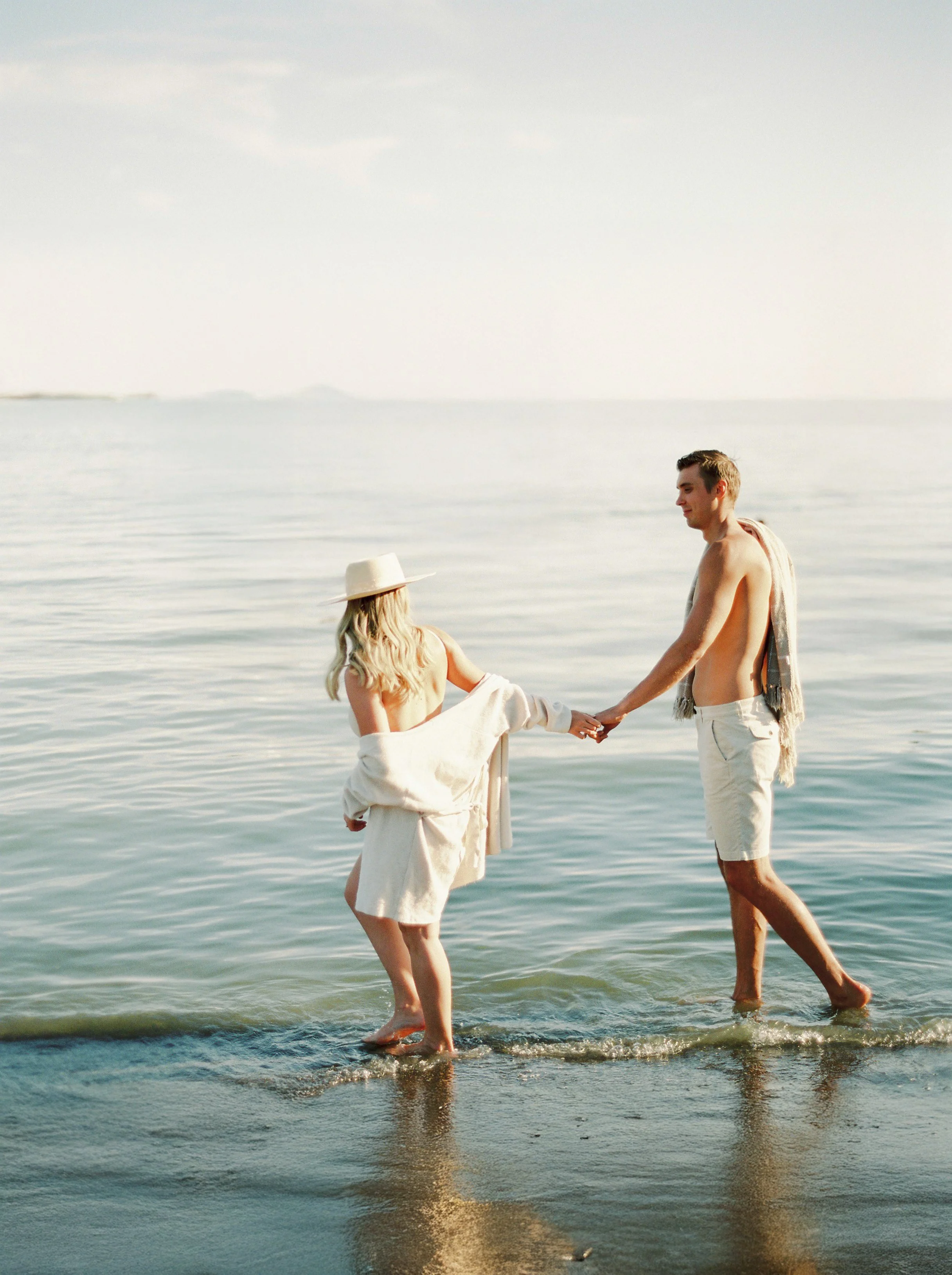 A man and woman holding hands and wading in the ocean at sunset, dressed in beachwear with the woman wearing a sunhat, the man has a towel over his shoulder.