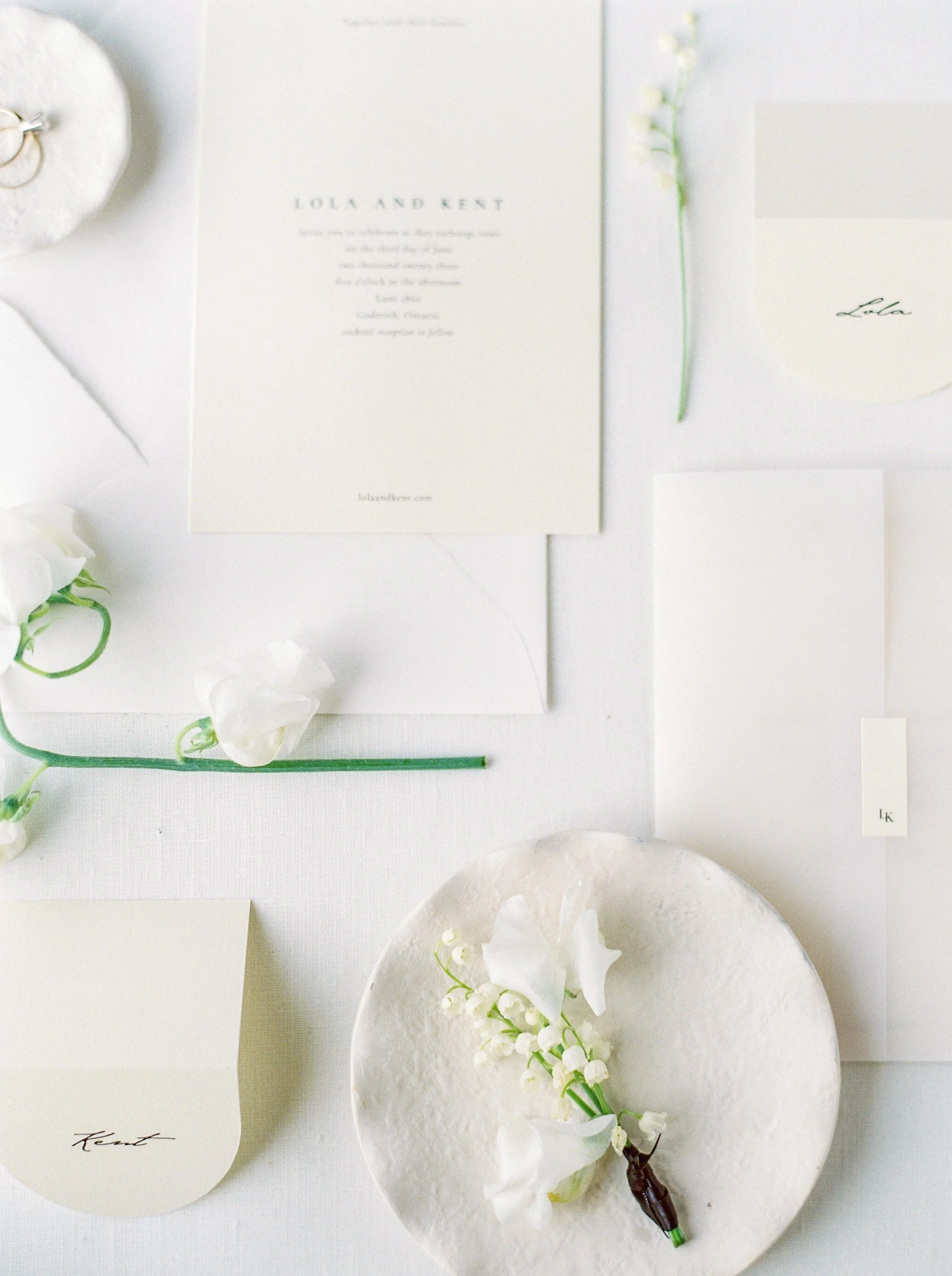 Flat lay of wedding stationery, white flowers, and a small white dish with flowers, all arranged on a white surface.