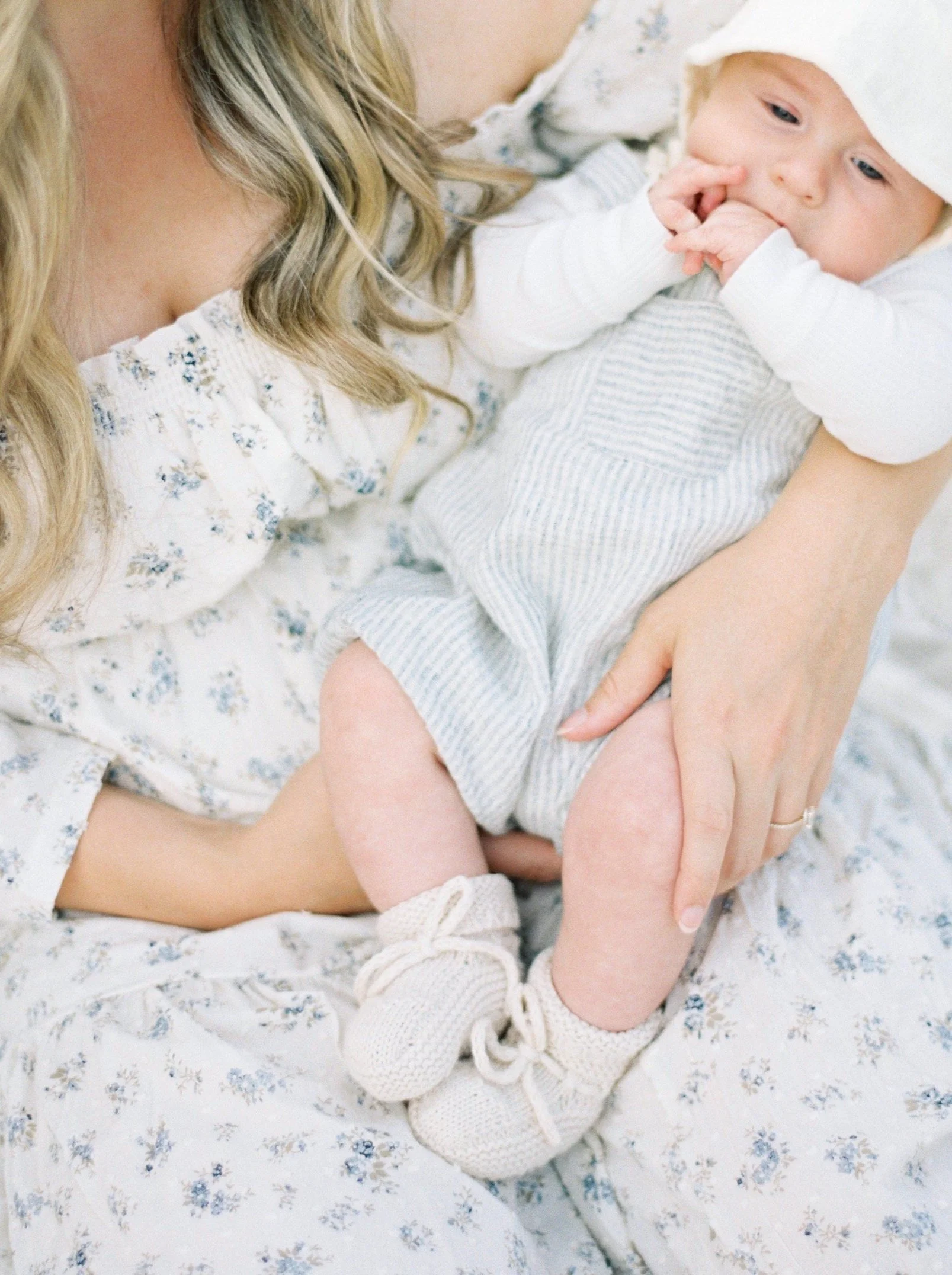 A woman holding a baby close, both dressed in white and light blue, with soft lighting and floral-printed bedding.