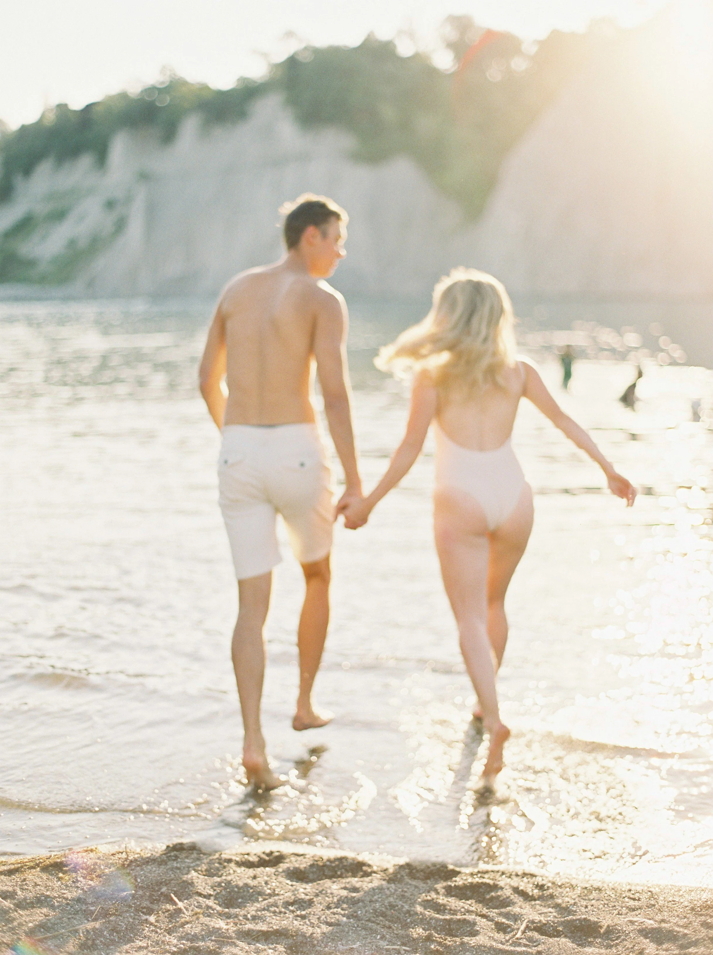 A young couple holding hands and walking into the water at the beach during sunset, with a rocky cliff in the background.