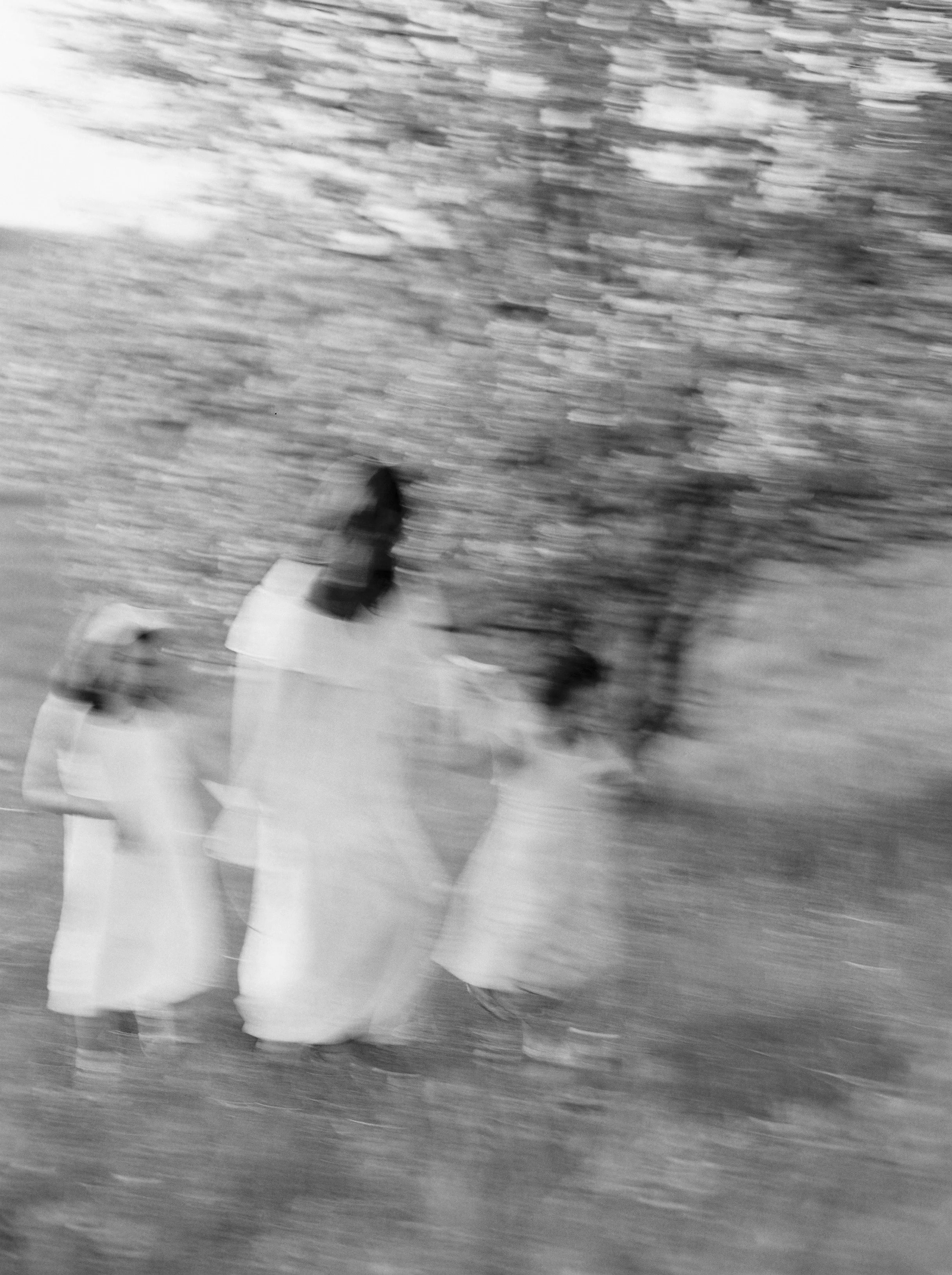 A blurry black-and-white photo of a group of three women walking outdoors near trees, dressed in long skirts, possibly on a farm or rural area.