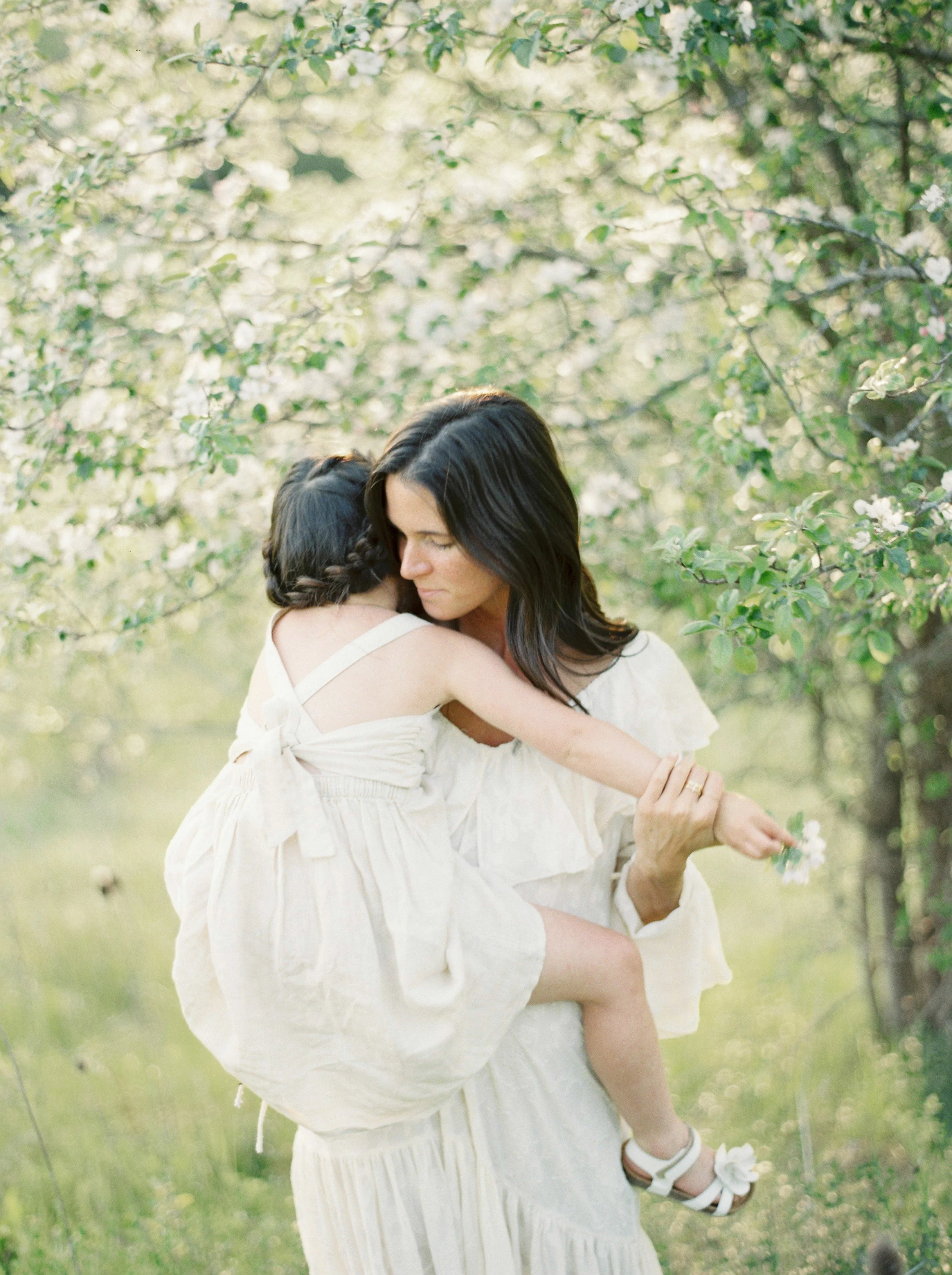 A woman carrying a child in a white dress through a green, flowering orchard, holding a small flower in her hand.