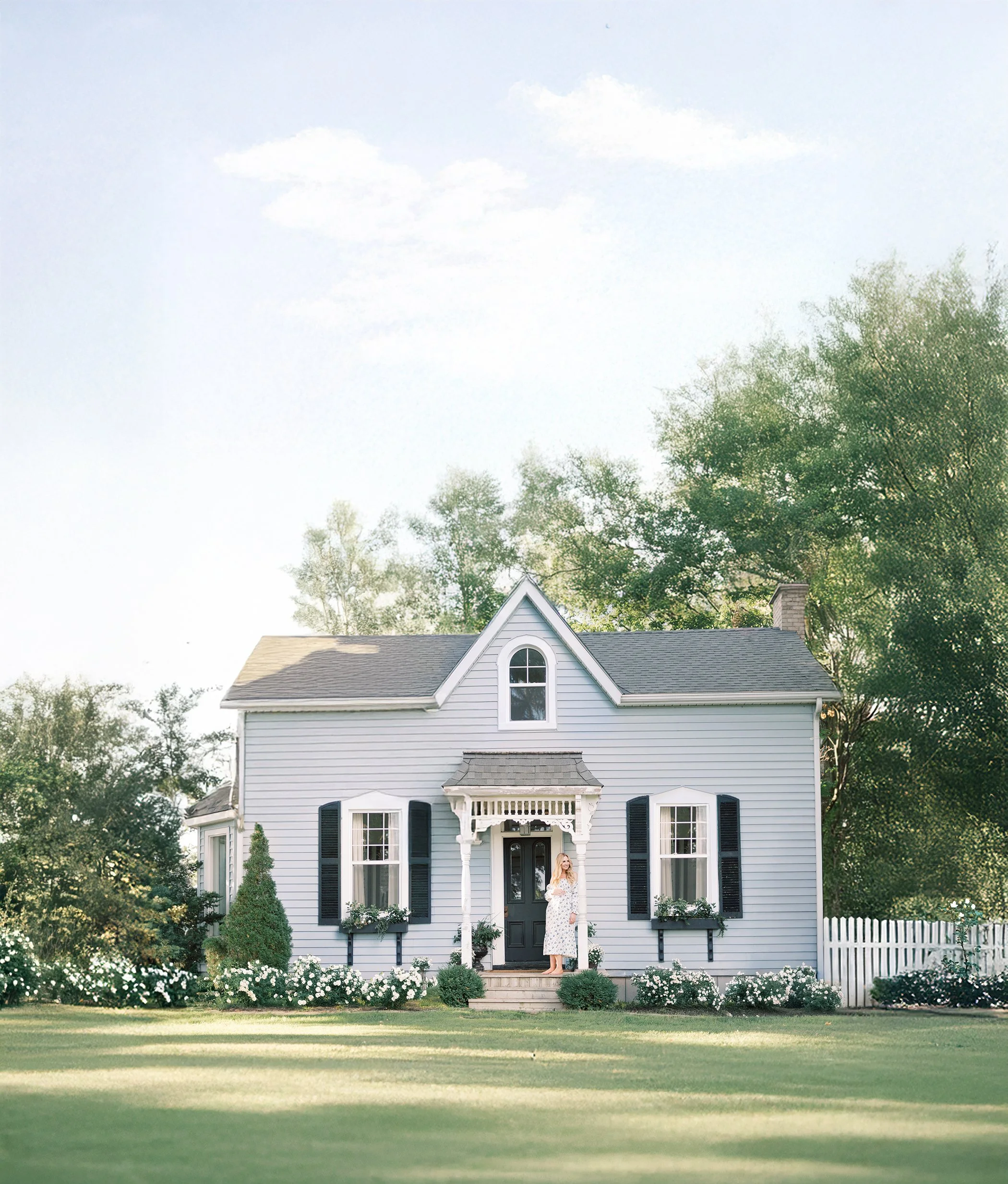 A woman in a white dress standing on the front steps of a two-story, light blue house with black shutters, surrounded by a well-maintained lawn and garden, with trees in the background and a partly cloudy sky.