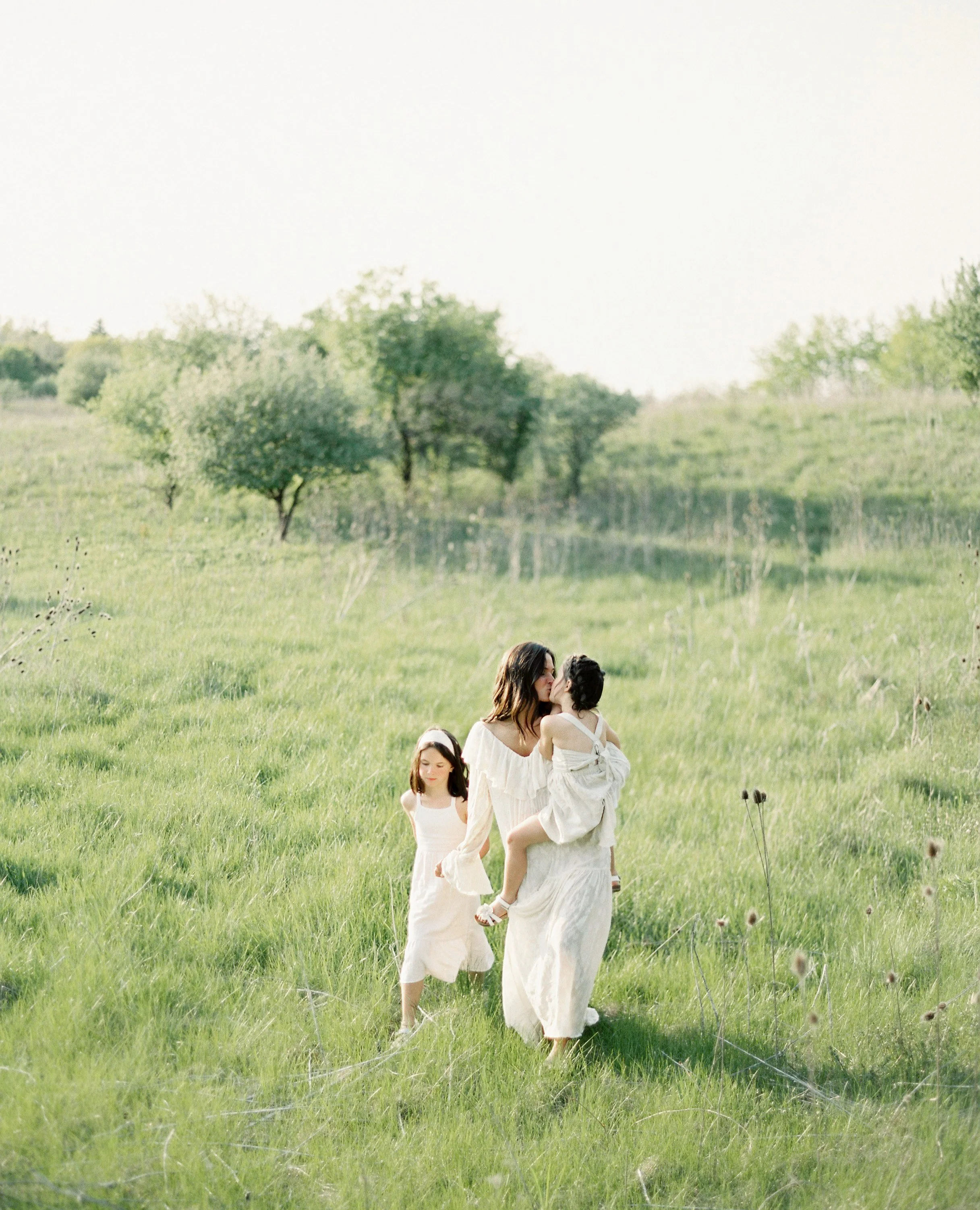 A woman and two girls walking through a grassy field with trees in the background, one girl is being carried and kissing the woman.