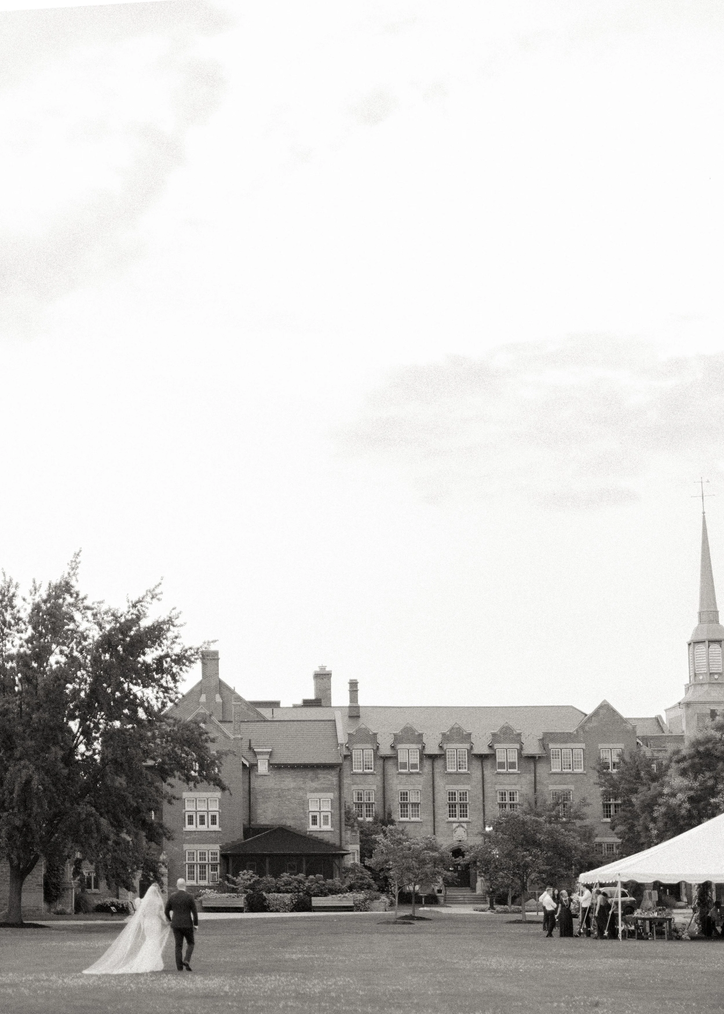 A bride and groom holding hands walking across a grassy area in front of a large historic building with multiple windows and a tall church steeple, during an outdoor wedding.