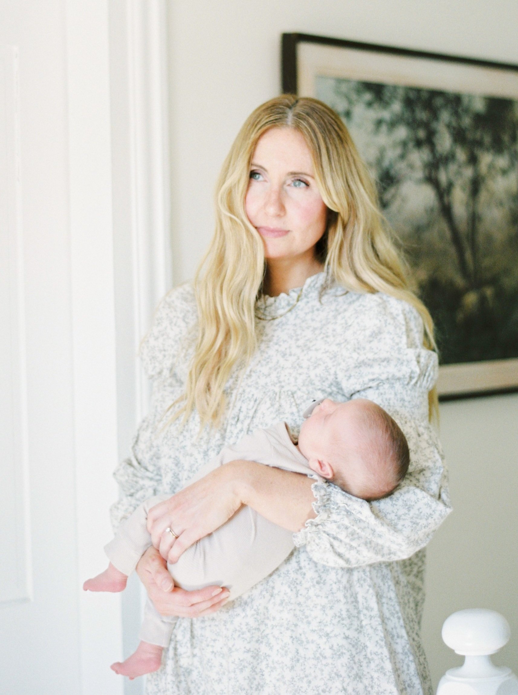 A woman with long blonde hair holding a sleeping baby, standing in a well-lit room with a painting on the wall.