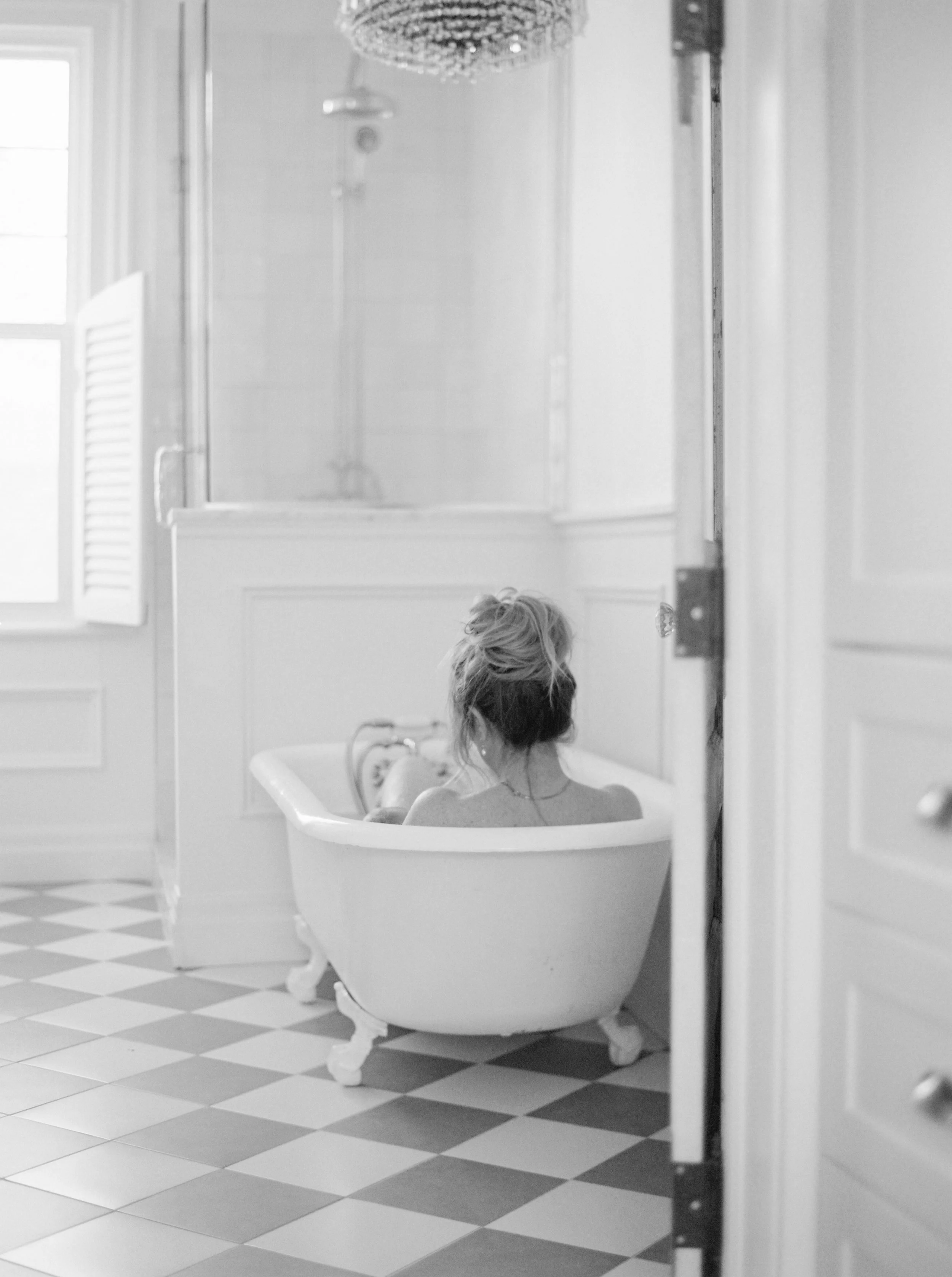 Black and white photo of a woman with messy hair sitting in a vintage clawfoot bathtub in a bright bathroom. The view is from outside the bathroom door, showing a checkered floor and a window with blinds on the wall.