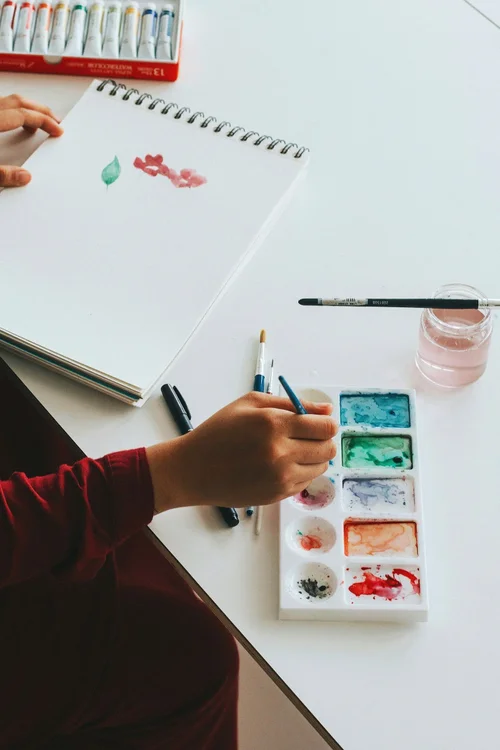 A person painting in a sketchbook with watercolor materials on a table
