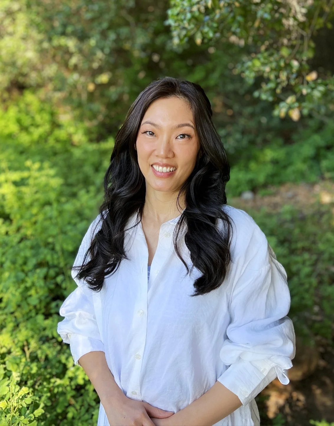 A person with long black hair wearing a white shirt, standing outdoors with greenery in the background.