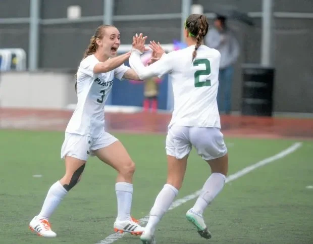 Two female soccer players wearing white uniforms celebrating on the field