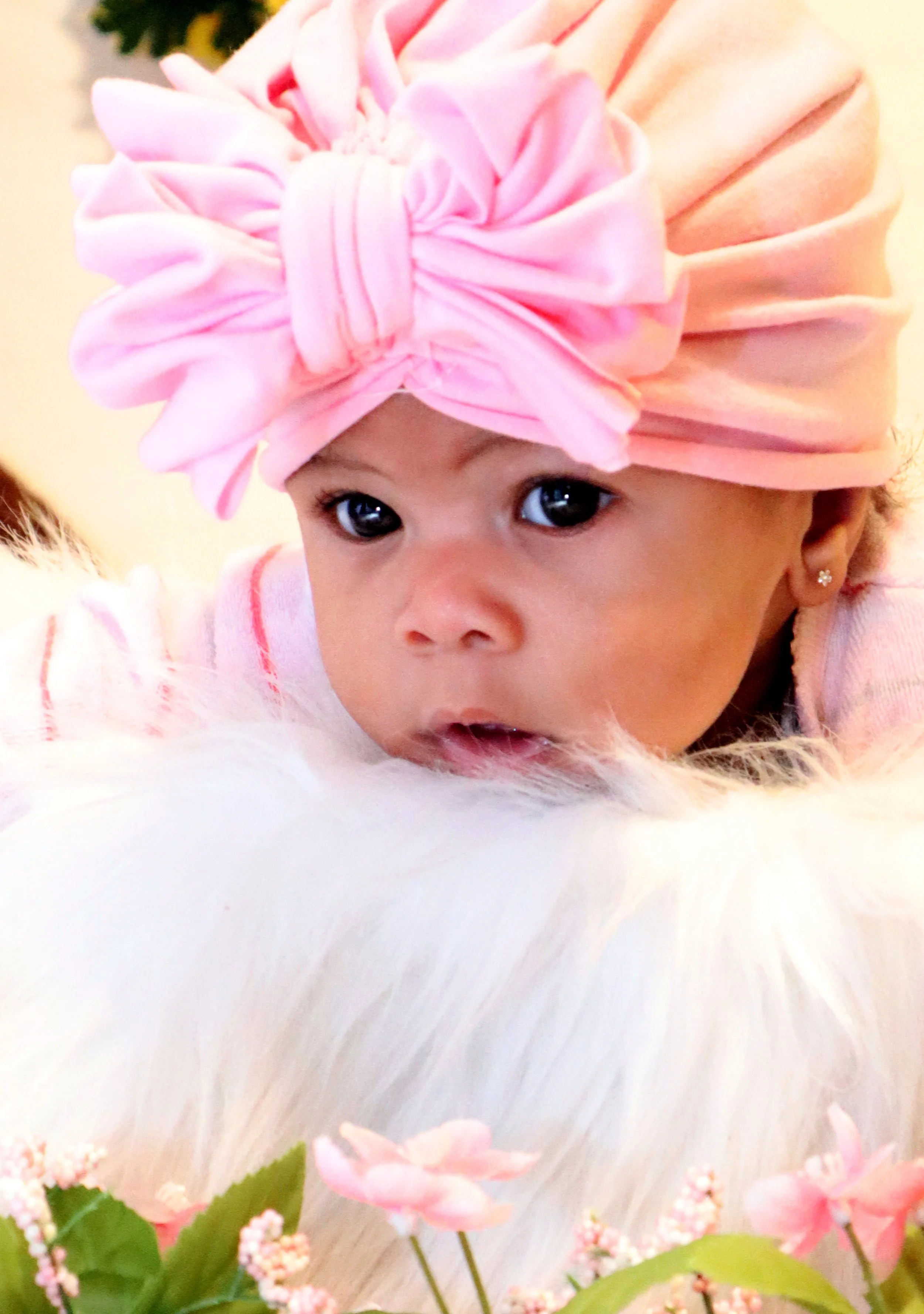 Baby wearing a pink headwrap with a bow, lying on a white fluffy surface with pink flowers in the foreground.