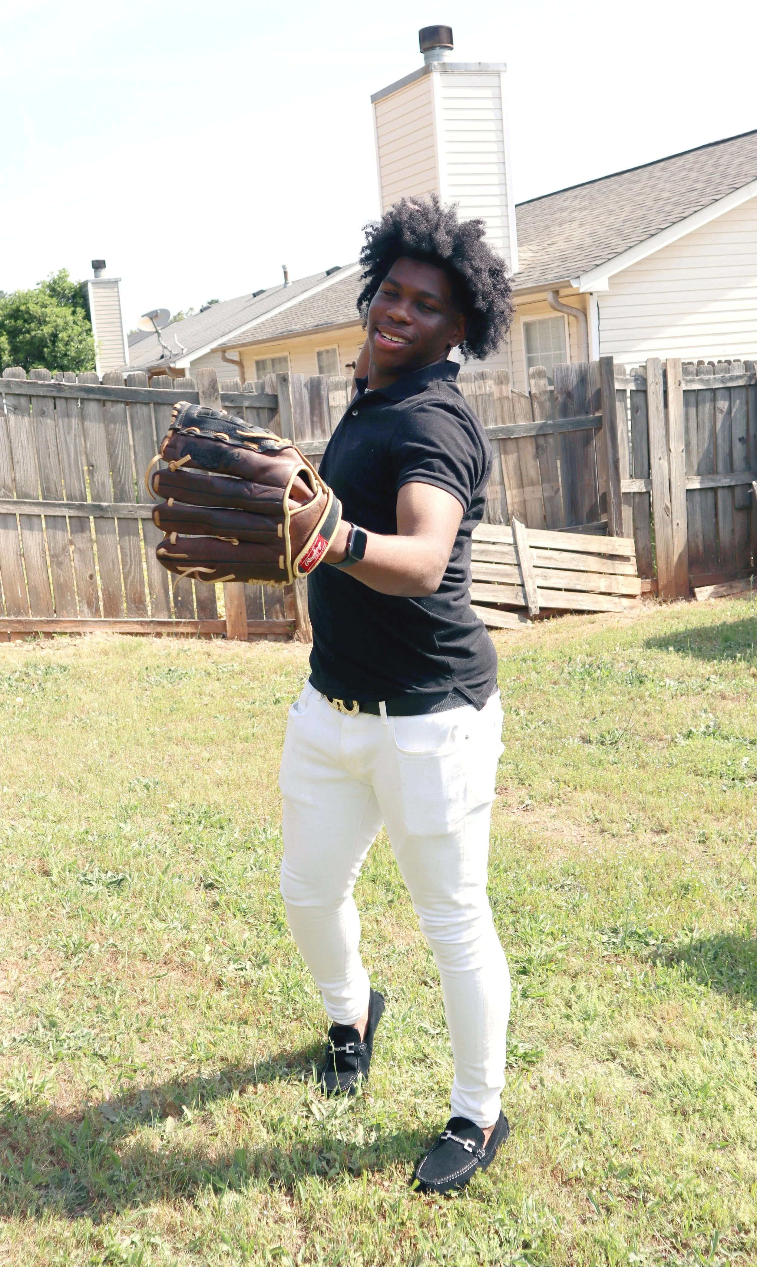 Person wearing a black shirt and white pants in a backyard, holding a baseball glove.