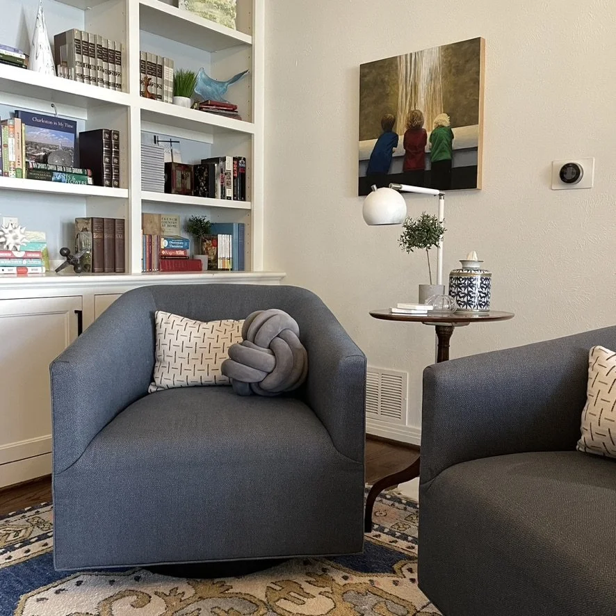Cozy living room with two gray armchairs, each featuring decorative pillows. A small round table holds a decorative lamp and jar. A bookshelf in the background contains books and decorative items, and a framed picture hangs on the wall.