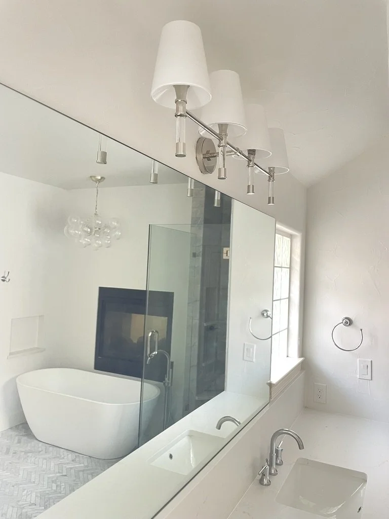 Modern bathroom with a large mirror and dual sink setup, featuring a white freestanding bathtub, glass shower enclosure, overhead light fixture, and a wall-mounted towel ring.