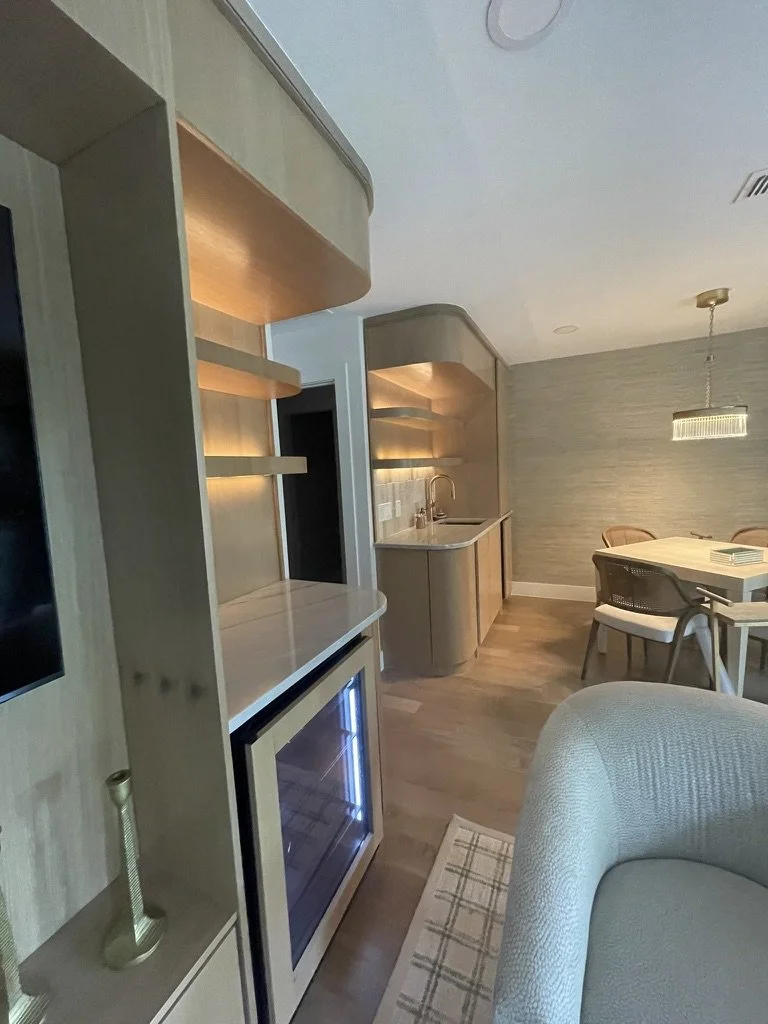 Modern kitchen and dining area with light wood cabinets, a small table with chairs, a wine cooler, and a cushioned armchair in the foreground.