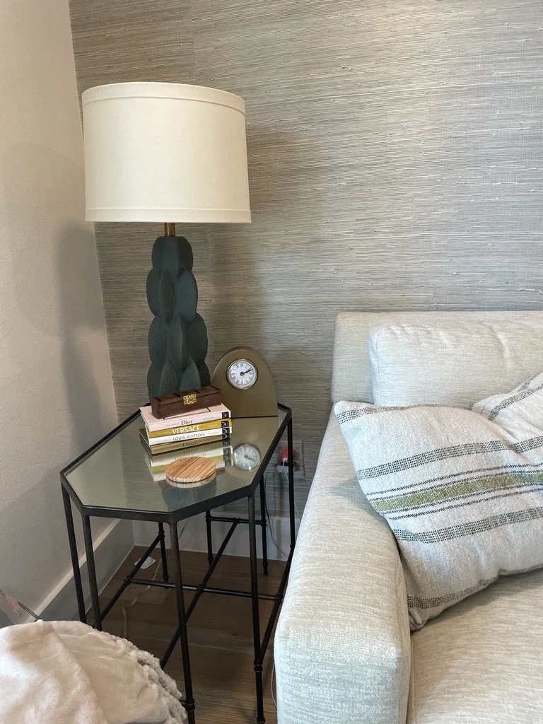 Living room corner with a modern glass side table, a decorative lamp, stacked books, a small clock, and a beige sofa with textured cushions against a light wood floor and neutral wall.