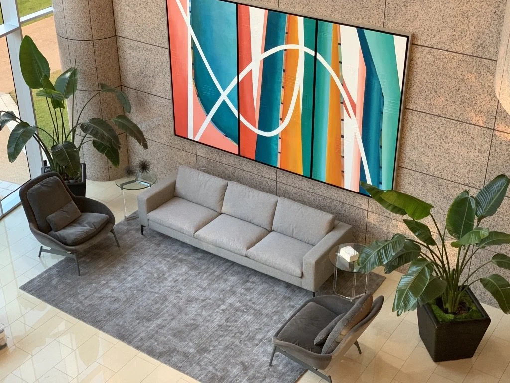Modern lobby with a gray sofa, two armchairs, large abstract painting, and indoor plants. The floor is tiled, and a grey rug is under the seating area.
