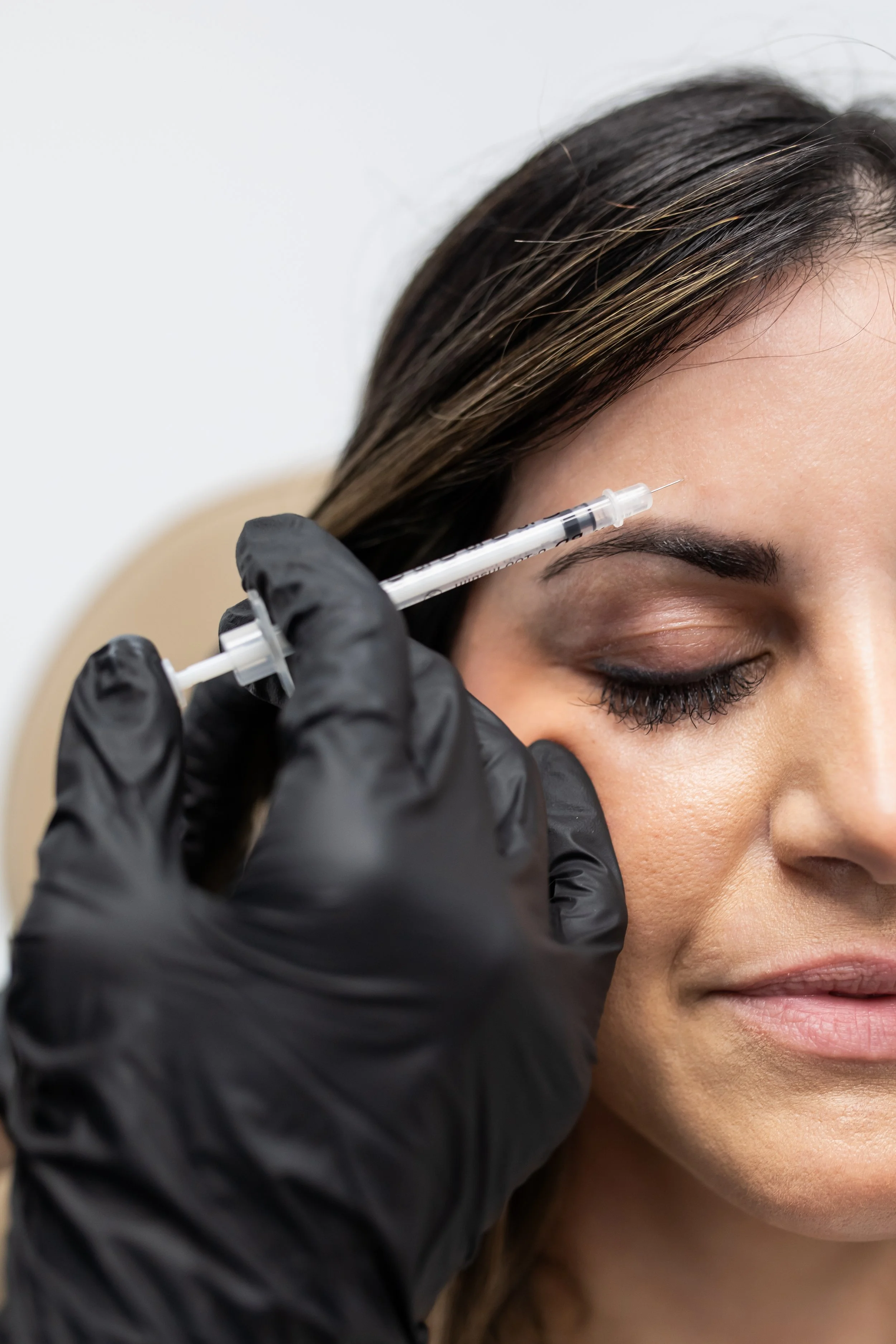 Close-up of a person receiving a cosmetic injection near the eyebrow by a practitioner wearing black gloves.