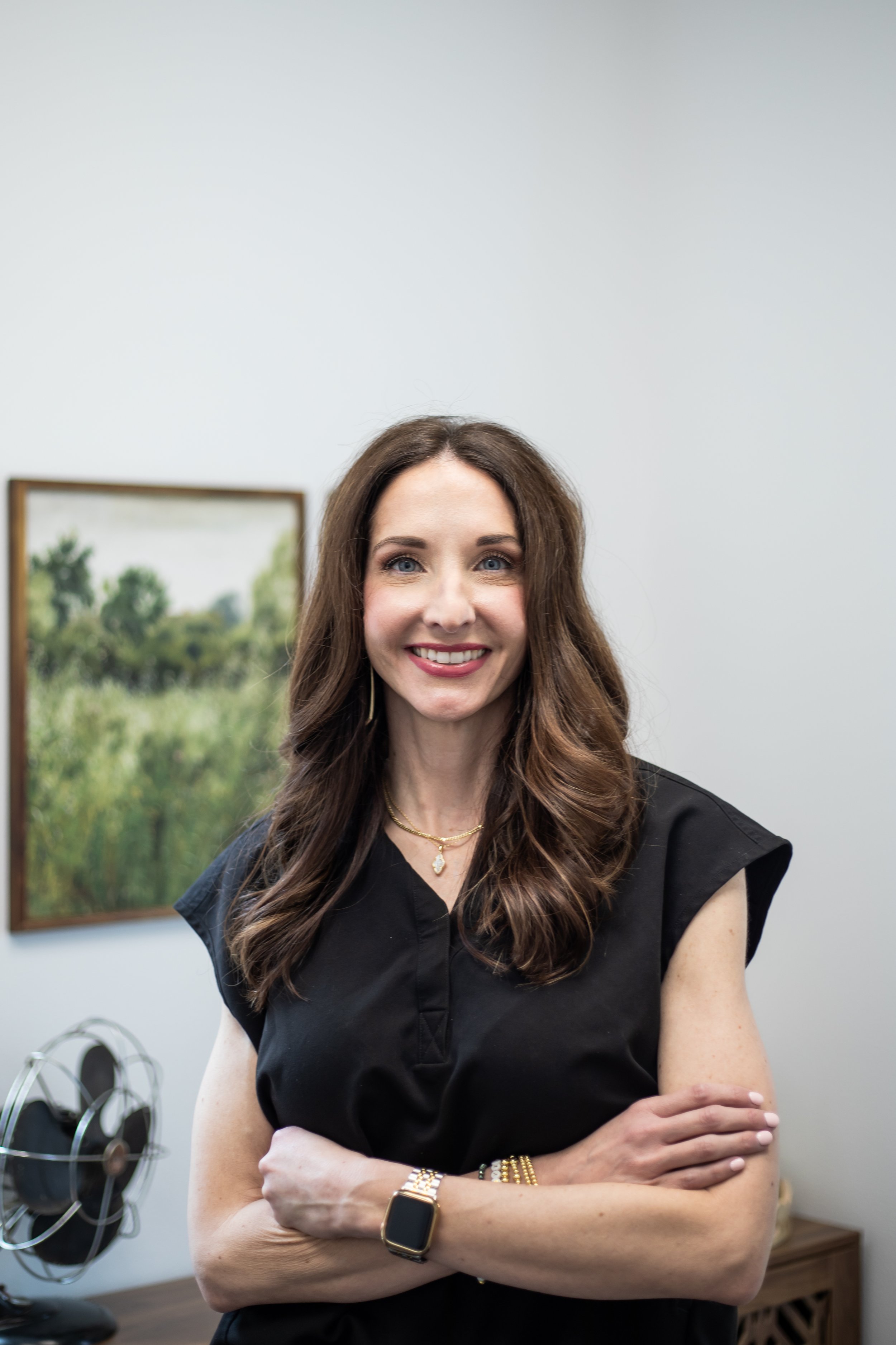 Woman with long brown hair and a black top smiling, standing in an office with a painting and a fan in the background.