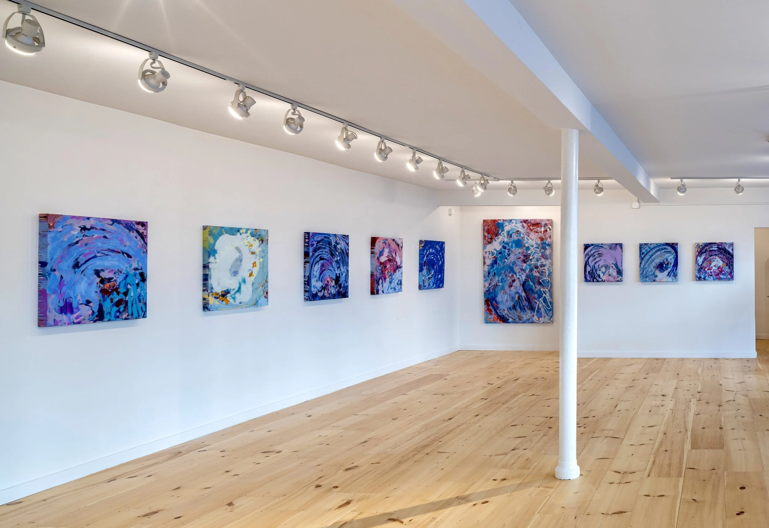Interior of an art gallery with abstract paintings on white walls, track lighting on the ceiling, and light hardwood flooring.