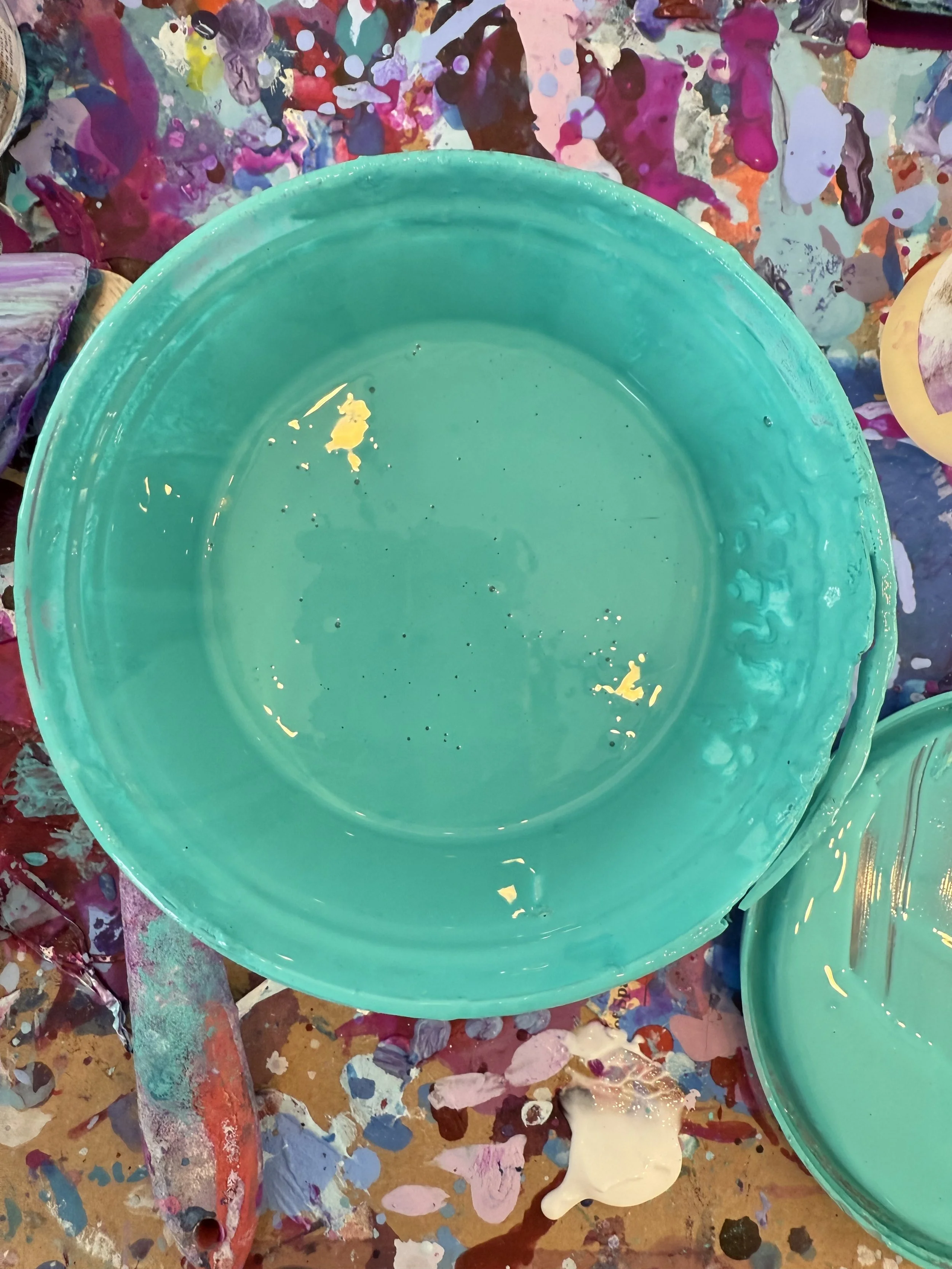 A large turquoise paint bucket with some yellow paint inside, placed on a colorful and messy artist's work surface splattered with various paints.