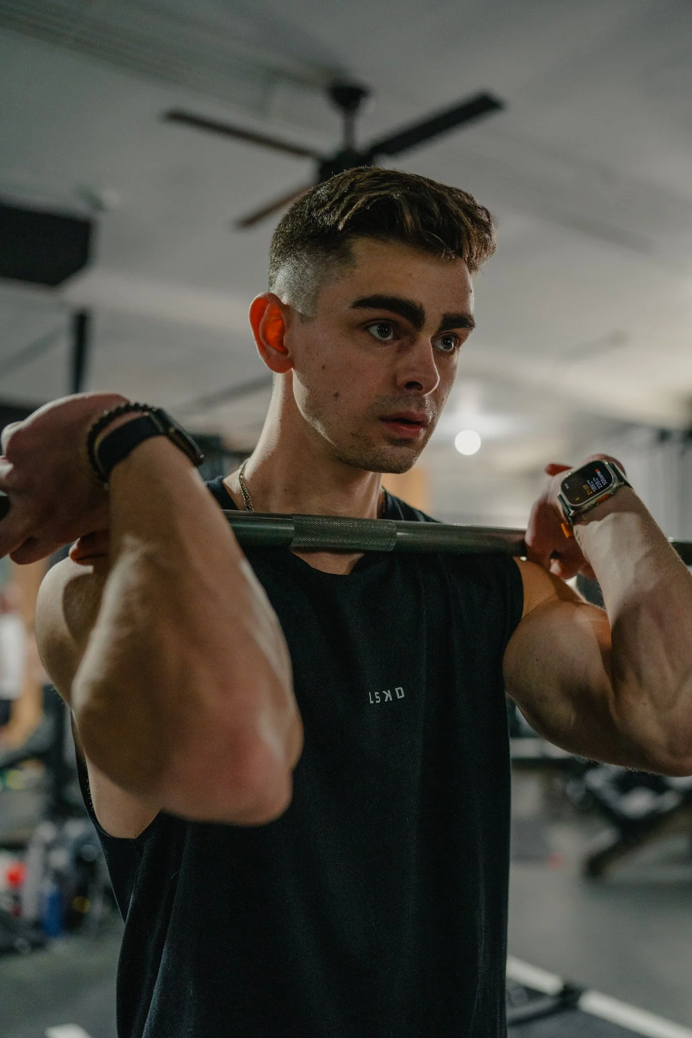 A young man in a gym lifting a barbell across his shoulders, wearing a digital smartwatch and a black sleeveless shirt, with a focused expression.