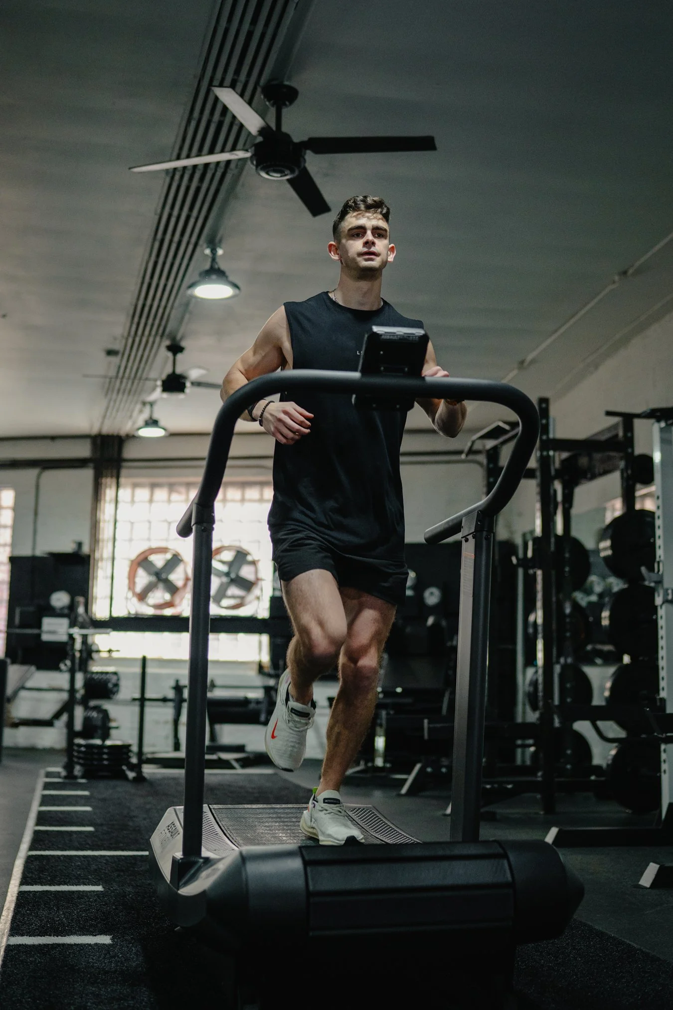 A man jogging on a treadmill in a gym, wearing a black sleeveless shirt and shorts.