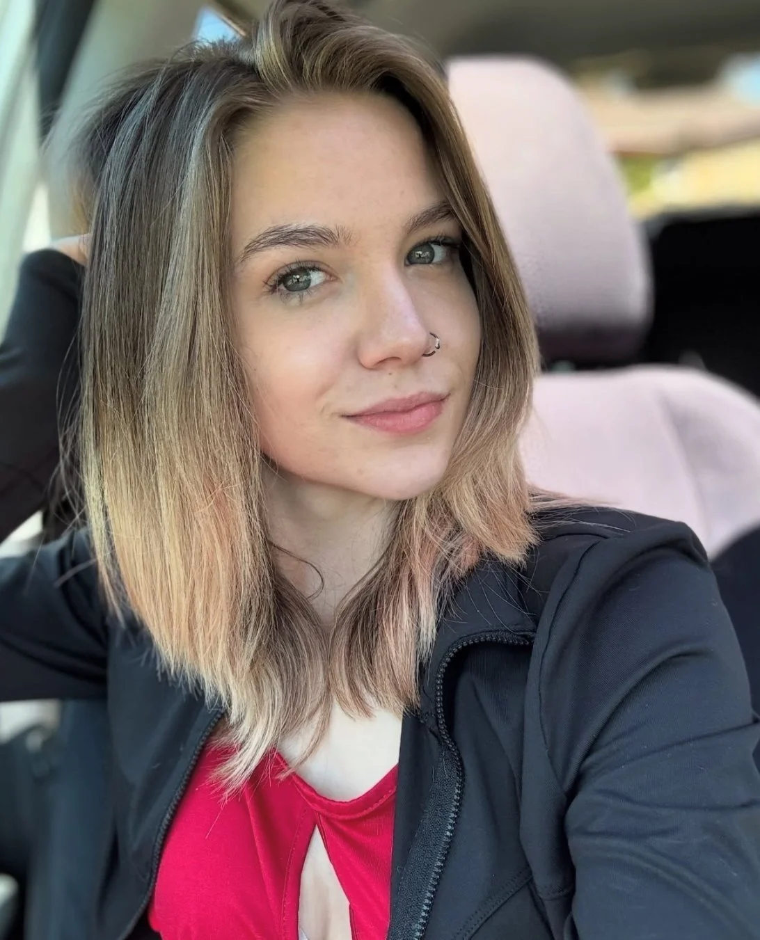 Person with shoulder-length hair and a nose ring wearing a red top and black jacket, sitting in a car.