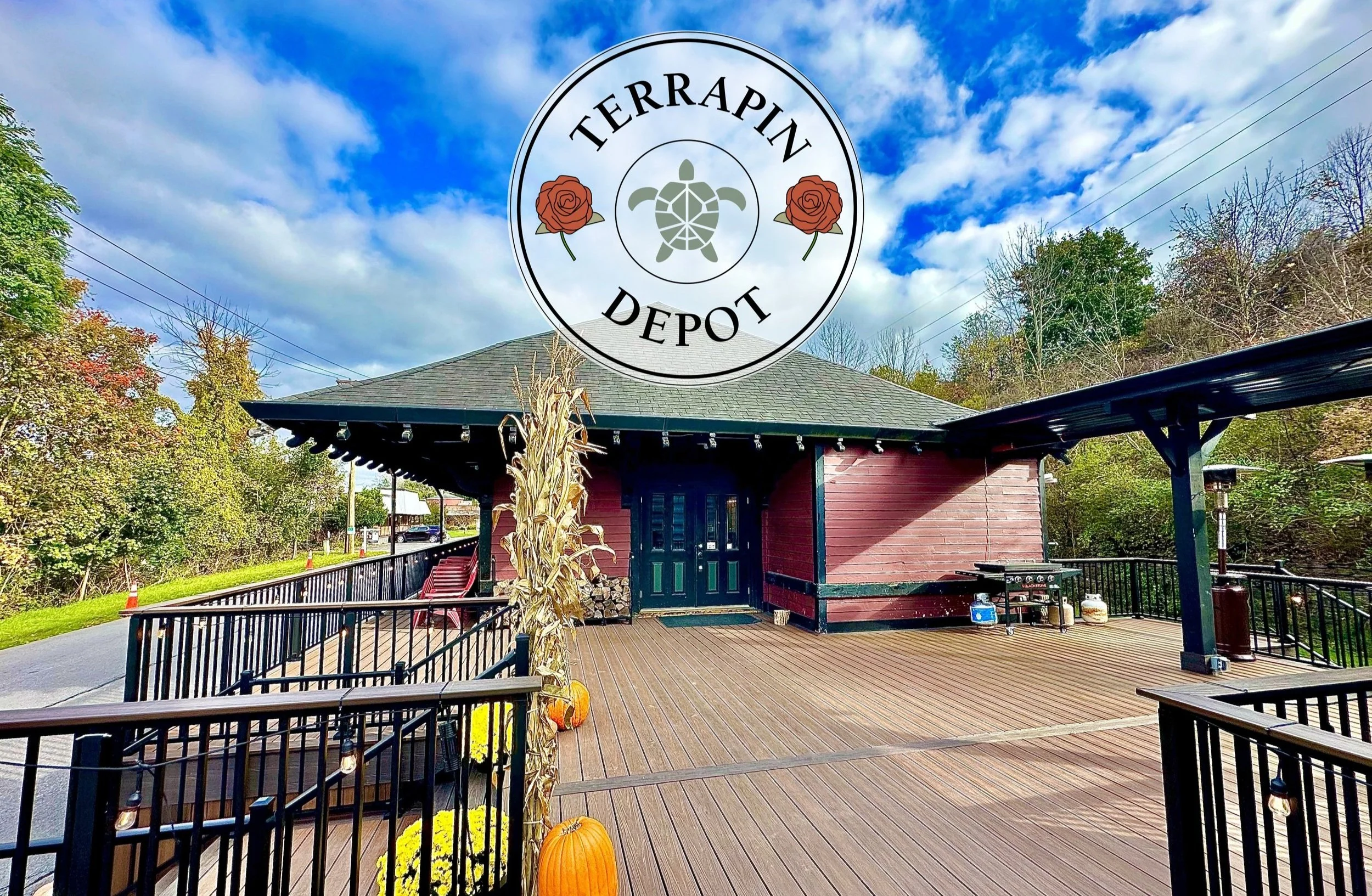 Historic train station building with "Terrapin Depot" logo, featuring a turtle and roses, a wooden deck with pumpkins, and surrounding trees under a blue sky.