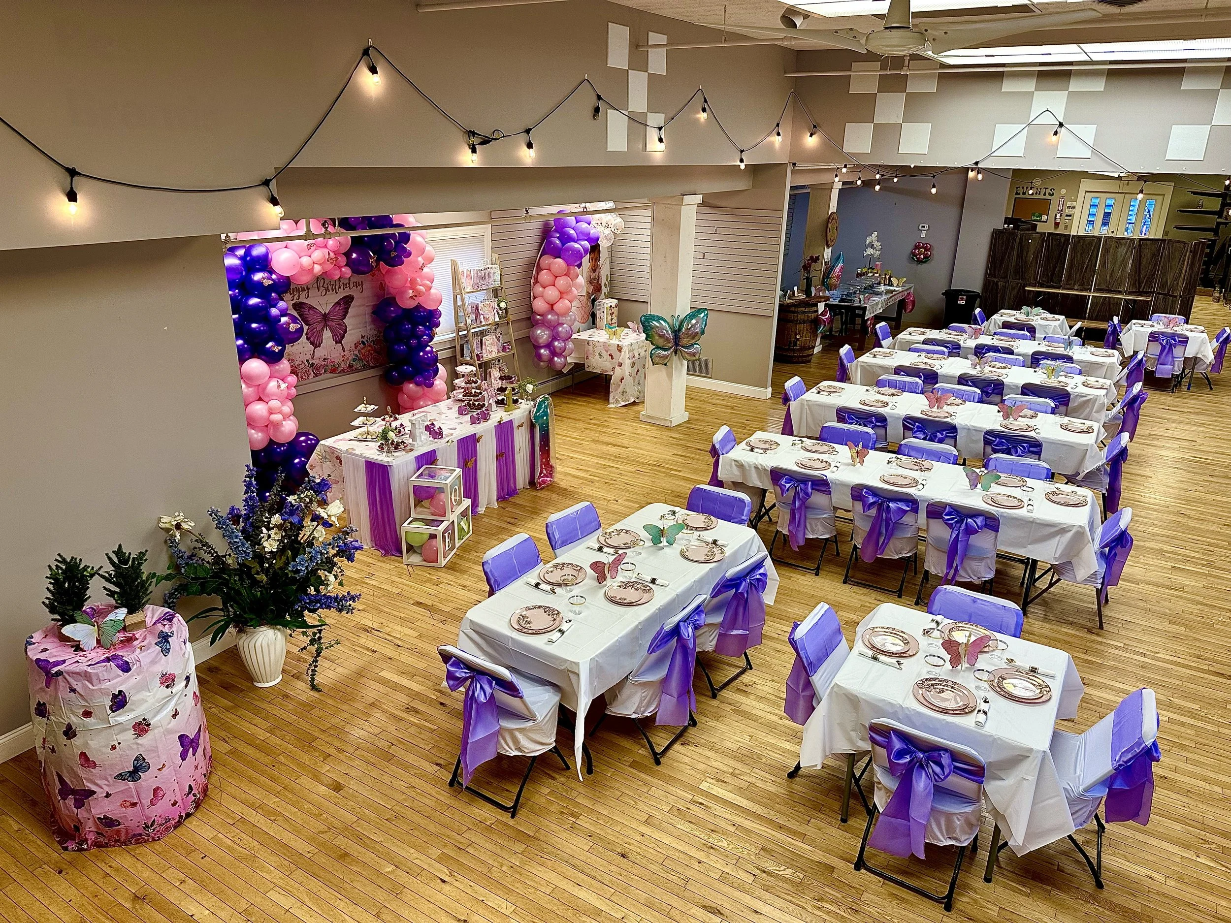 Event hall decorated for a birthday party with a butterfly theme. Tables are covered with white tablecloths and decorated with purple chair ribbons, butterfly-themed plates, and napkins. A backdrop with balloons and butterfly motifs is visible, along with string lights overhead.