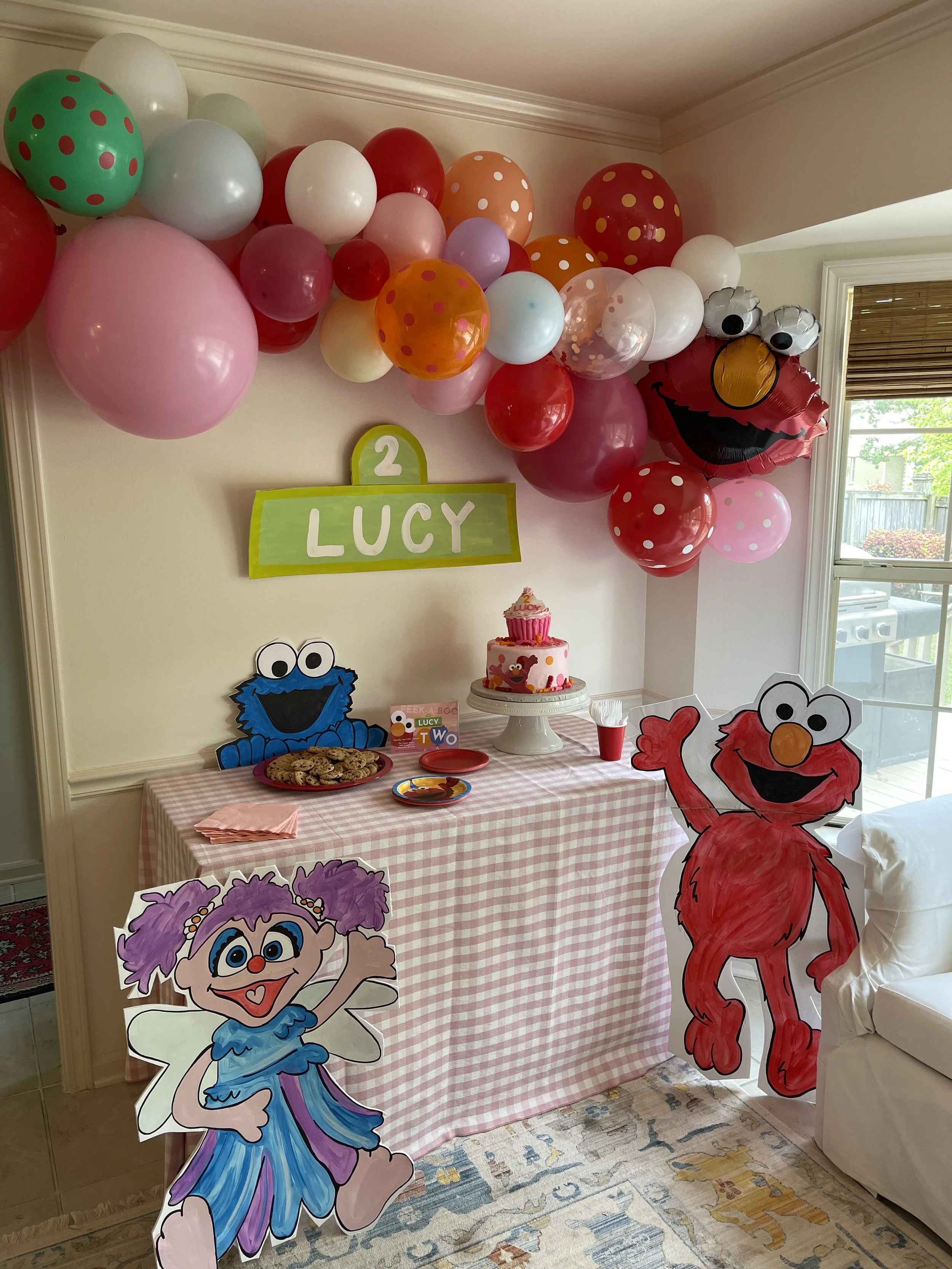 Children's birthday party setup with colorful balloons and cartoon character decorations of Elmo and other characters. A table with a pink checkered cloth holds a cake and cookies.