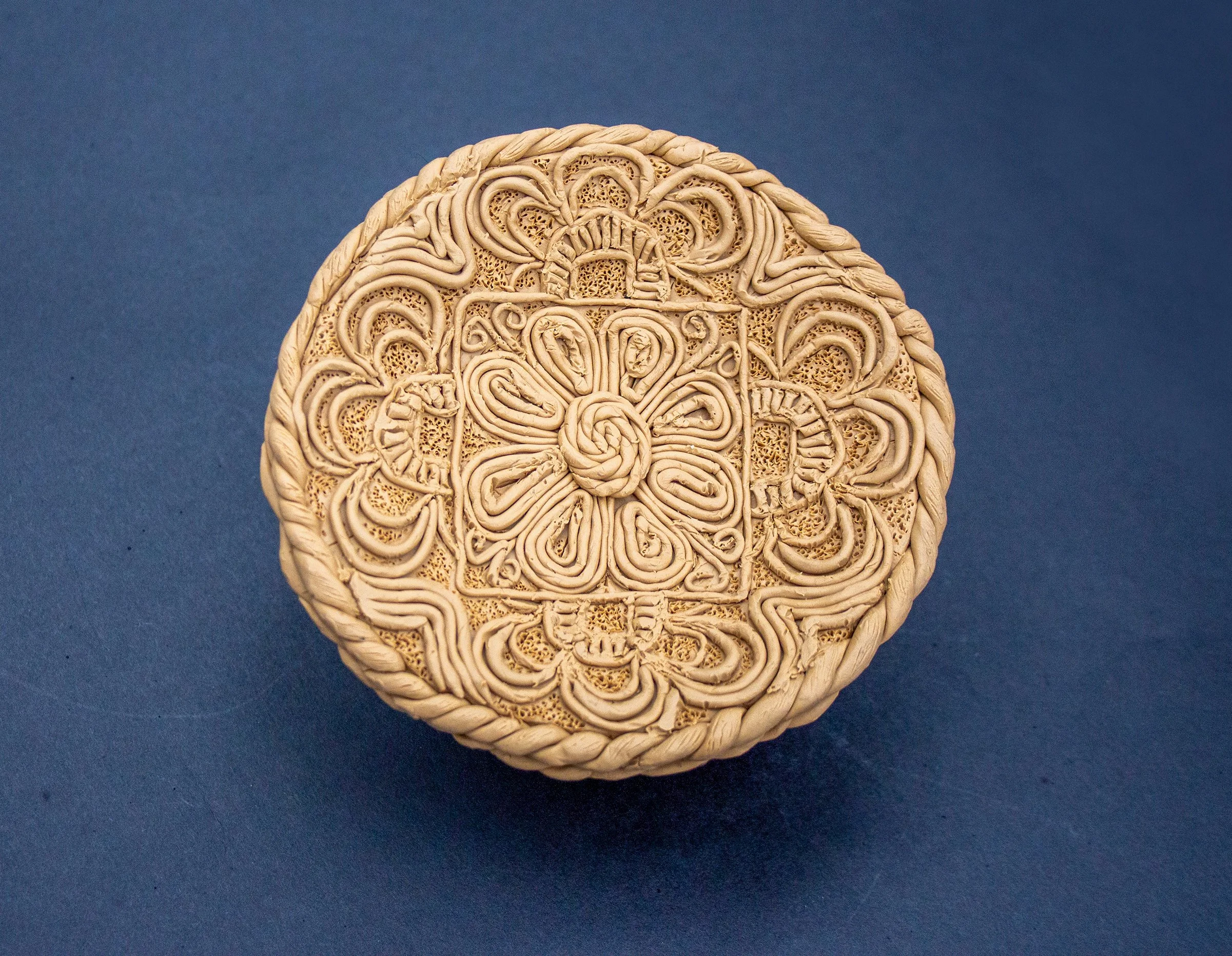Circular decorative item with intricate, symmetrical patterns and a braided edge, placed on a dark blue background.