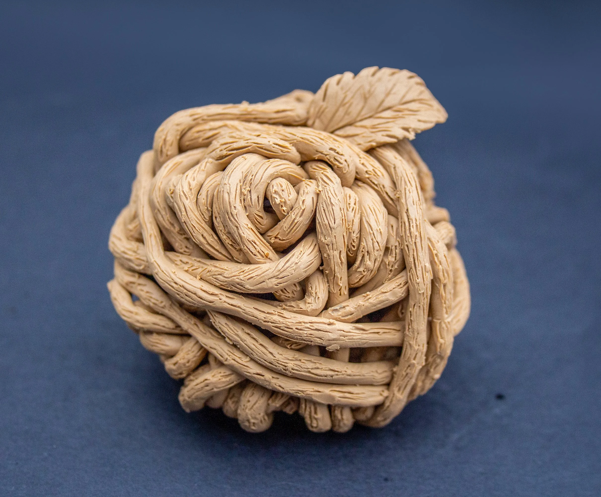 A beige, intricately woven ball made of rubber bands with a small rubber band leaf on top, placed on a blue surface.