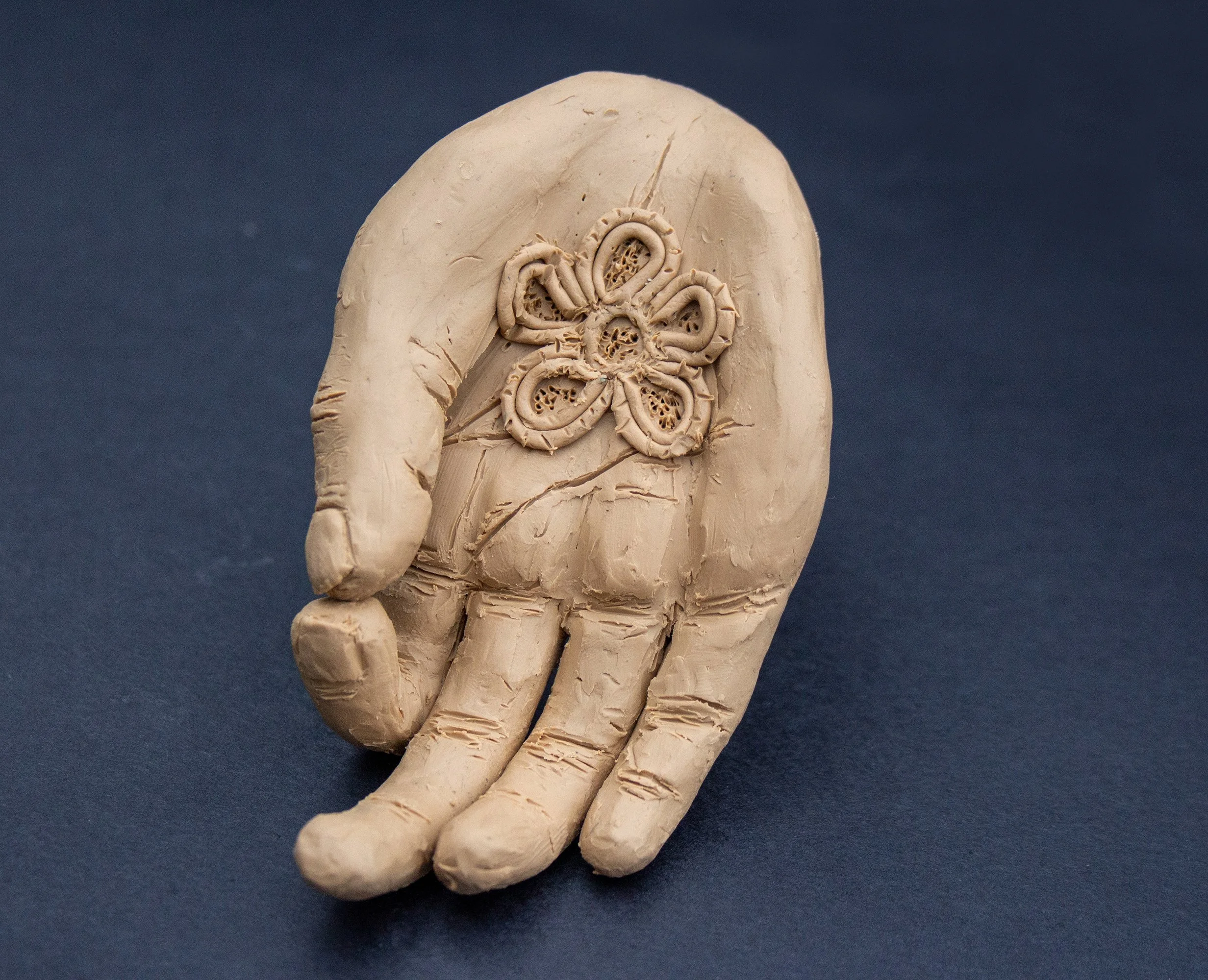 Ceramic sculpture of a hand holding a detailed flower with multiple petals, on a dark blue background.
