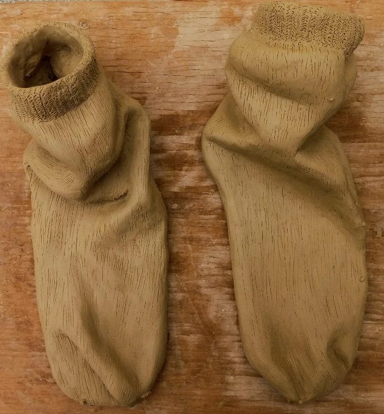 Two beige, clay or ceramic slipper-like objects with textured surface, placed on a wooden surface.