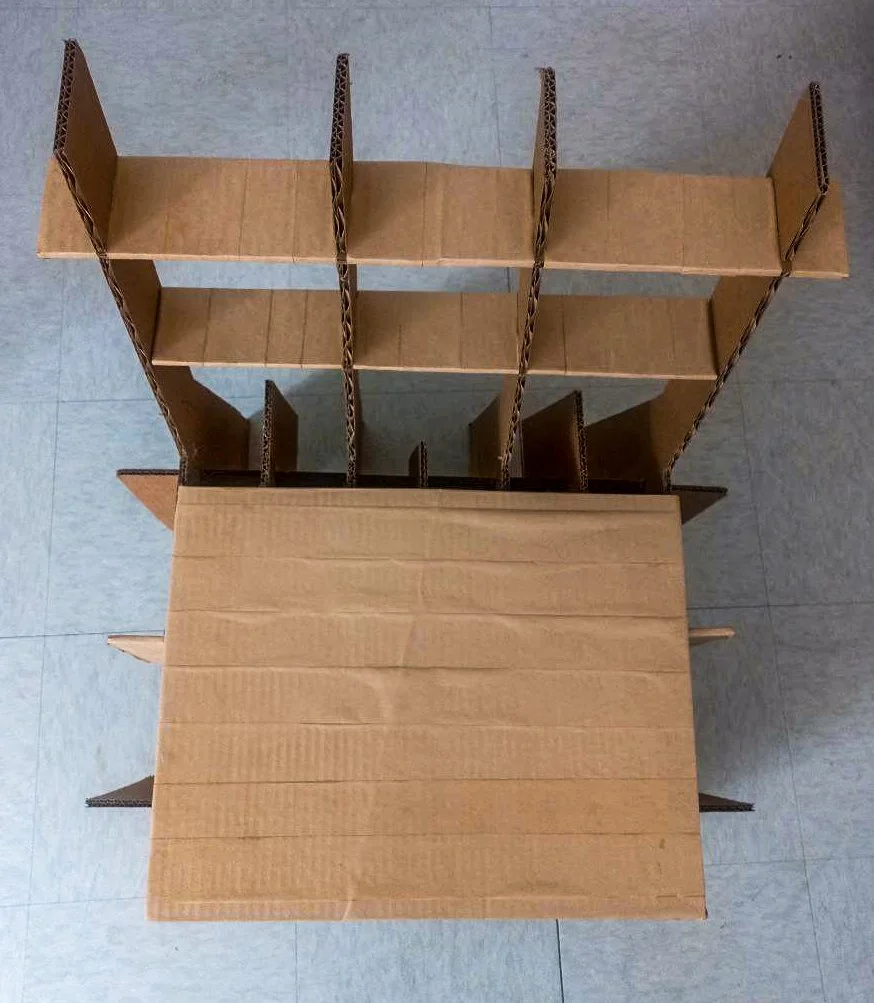 Overhead view of a partially assembled cardboard shelving unit with multiple compartments, placed on a tiled floor.