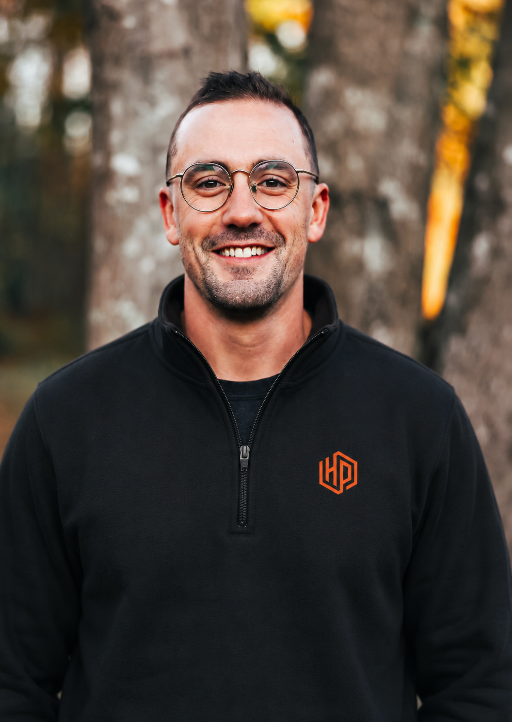 A smiling man with short dark hair, glasses, and a beard wearing a black jacket with an orange logo, standing outdoors near trees with autumn foliage in the background.